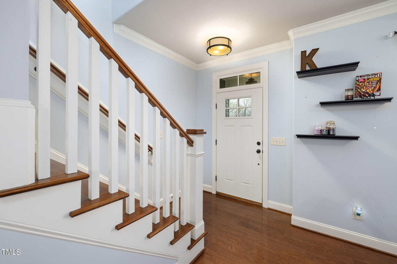 A brightly lit hallway features a wooden staircase with white railings, light blue walls, and a white front door with glass panels. On the right, floating shelves hold small items and a magazine. A ceiling light fixture illuminates the space.