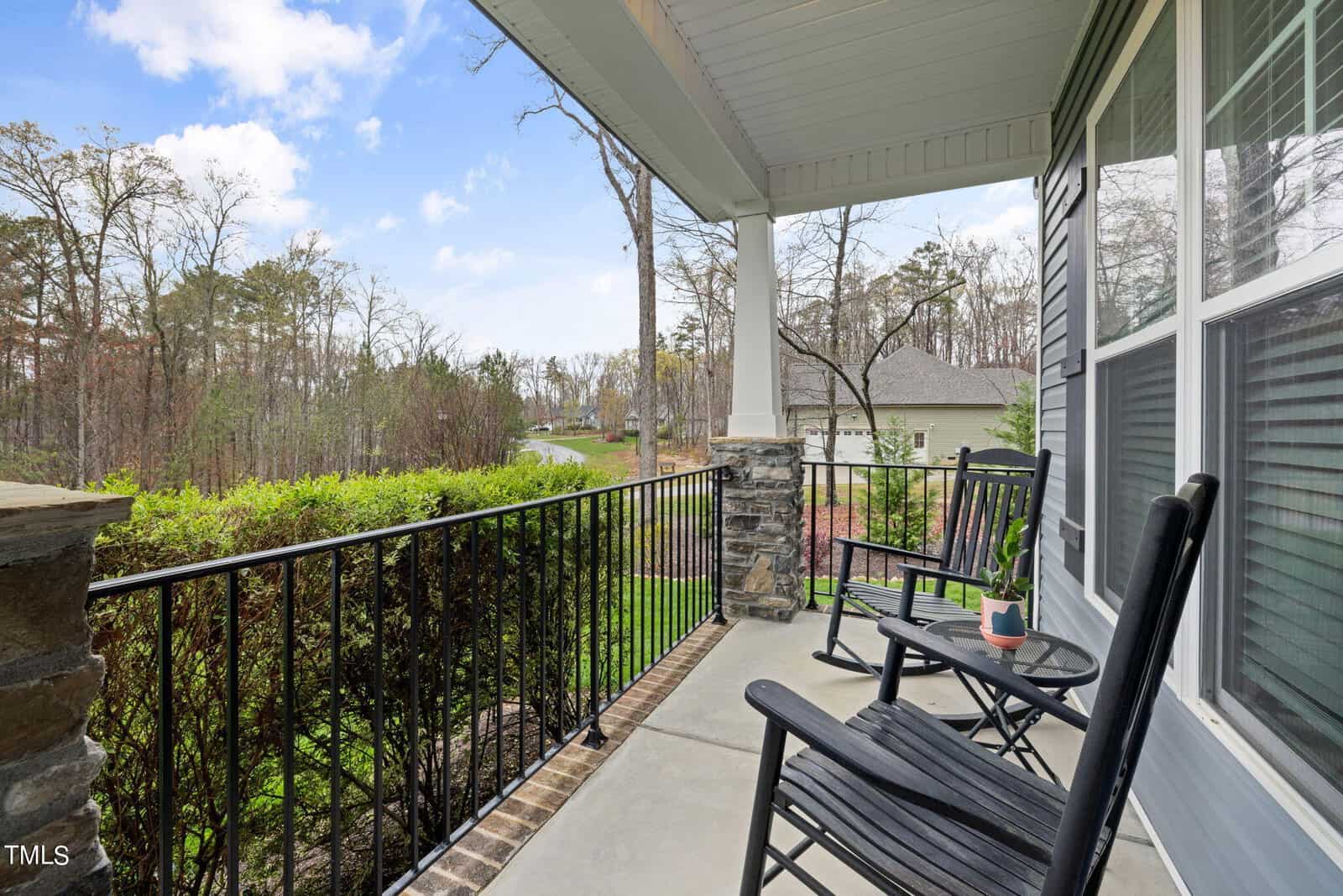 A cozy porch features two black rocking chairs and a small table with a potted plant. The porch overlooks a green lawn and trees under a clear blue sky with scattered clouds.