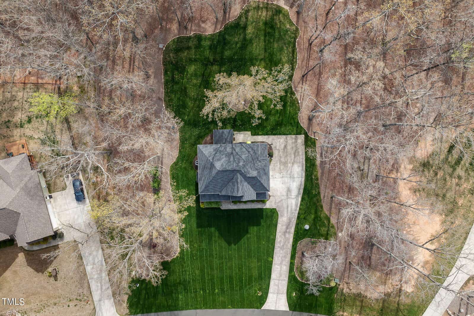 Aerial view of a house with a large green lawn, surrounded by leafless trees. The property has a curved driveway leading to the house, and neighboring homes are partially visible. The landscape appears to be in early spring.