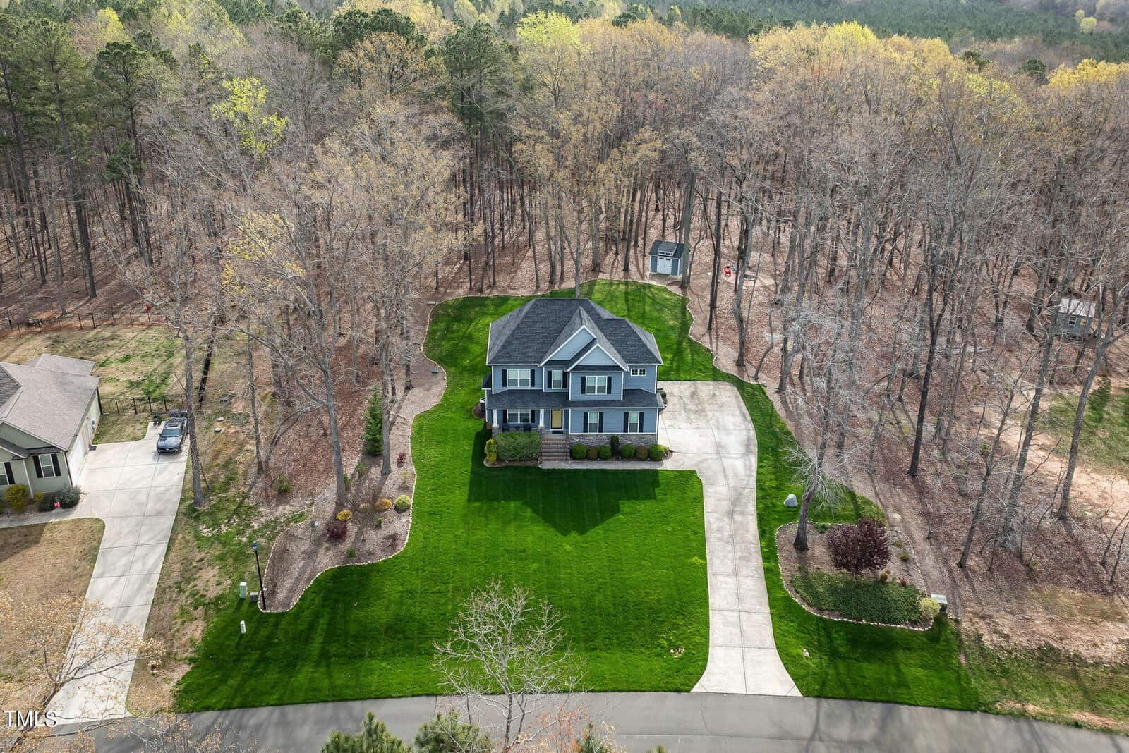 Aerial view of a two-story house with a gray facade, surrounded by a well-manicured lawn and bordered by tall, leafless trees. A curving driveway leads to the garage. The scene suggests a suburban neighborhood setting in early spring.