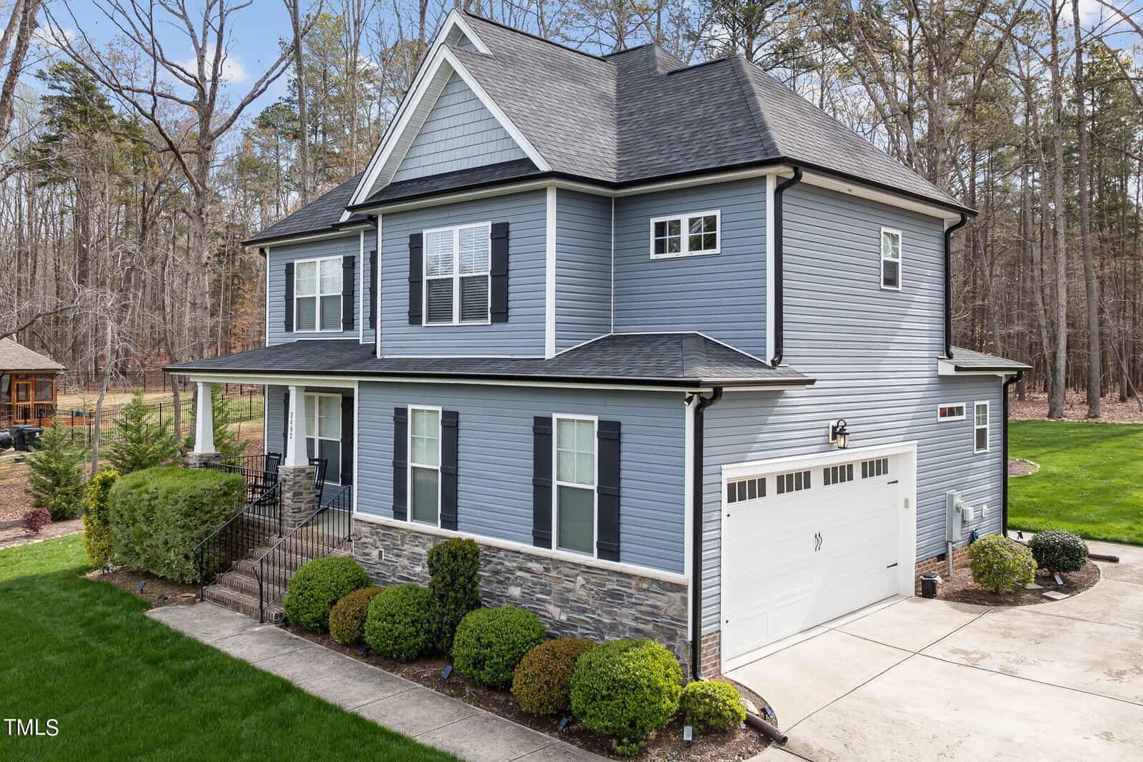 A two-story blue house with white trim and a shingled roof, featuring a garage. It's surrounded by shrubs and a well-maintained lawn, set against a backdrop of leafless trees on a clear day.