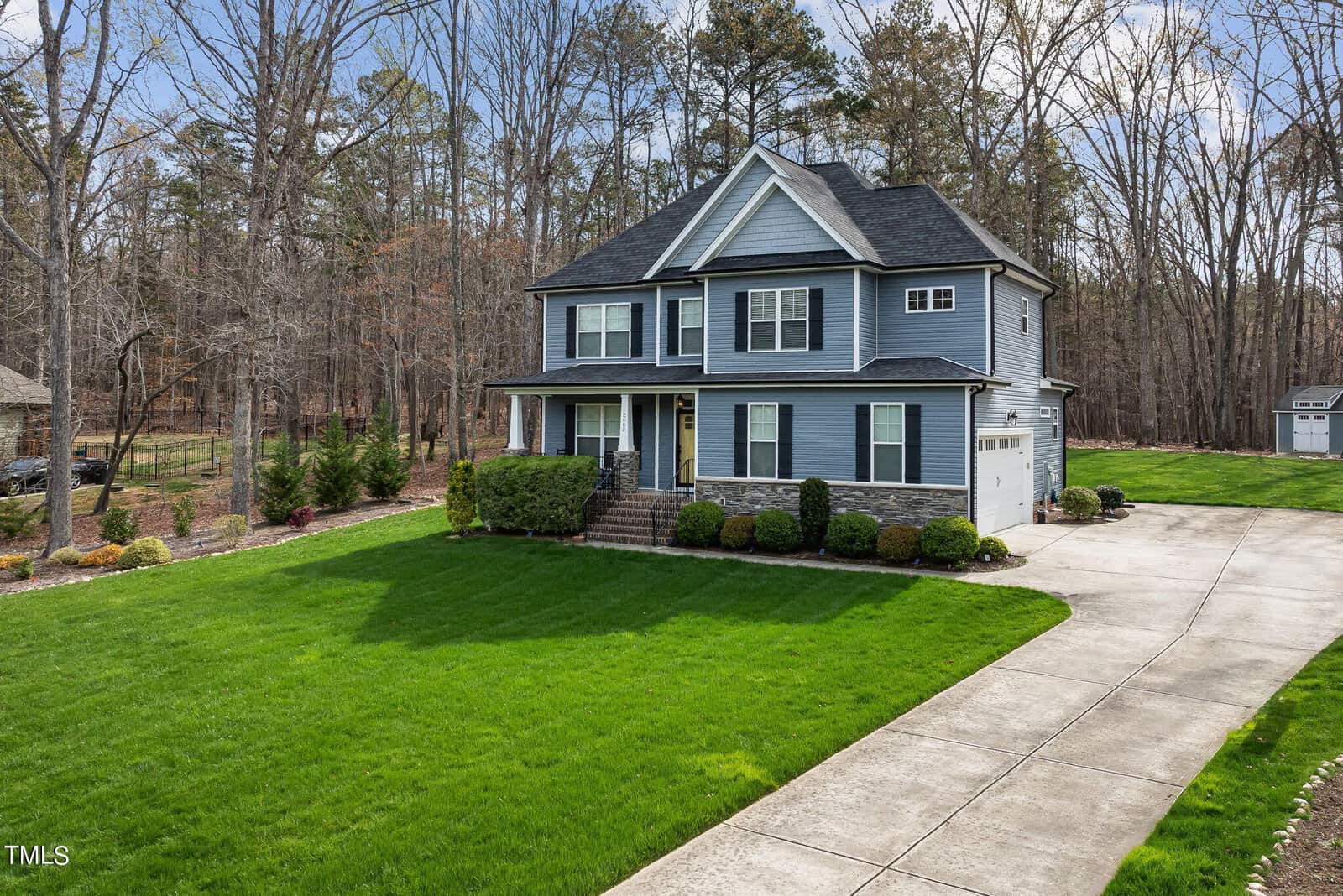 A two-story gray house with a dark roof, white trim, and a front porch. It's surrounded by neatly landscaped lawns and bushes, adjacent to a wooded area. A long driveway leads to a white garage. The sky is clear and sunny.