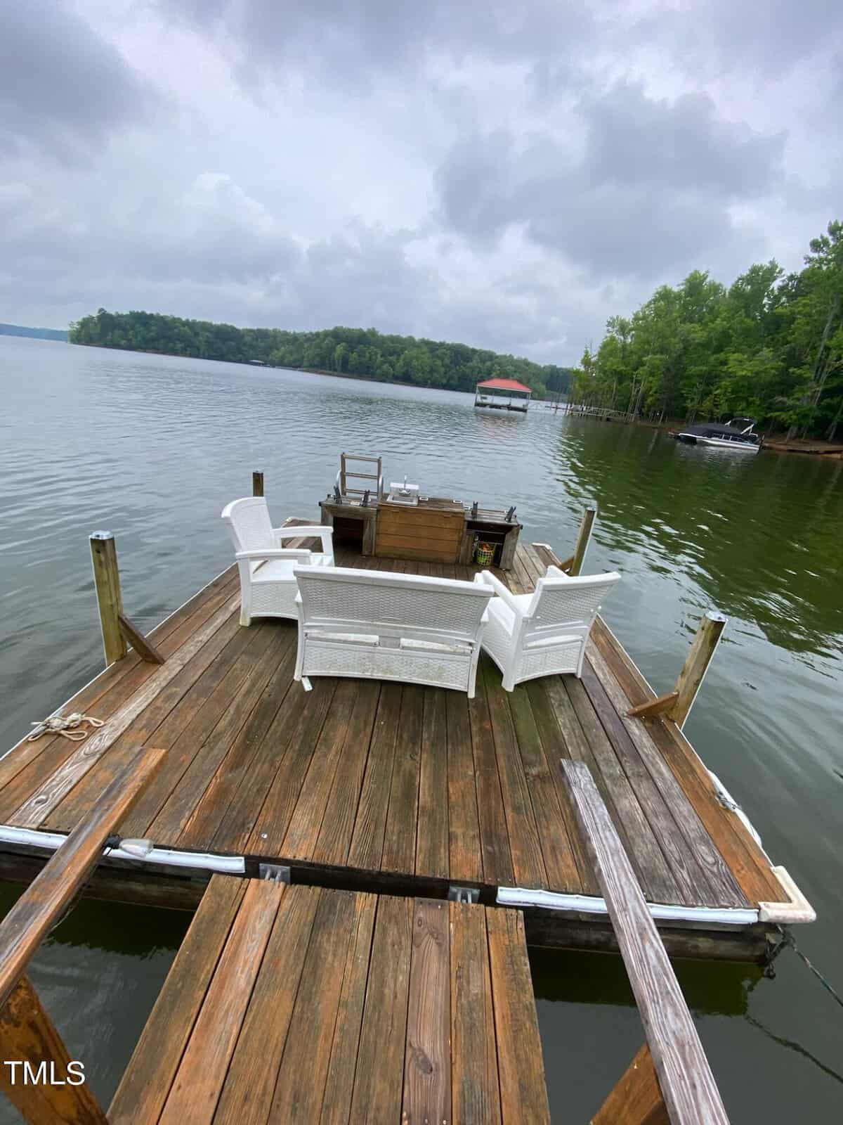 A serene lakeside wooden dock with white outdoor furniture and a small table, surrounded by trees, with boats nearby under a cloudy sky.