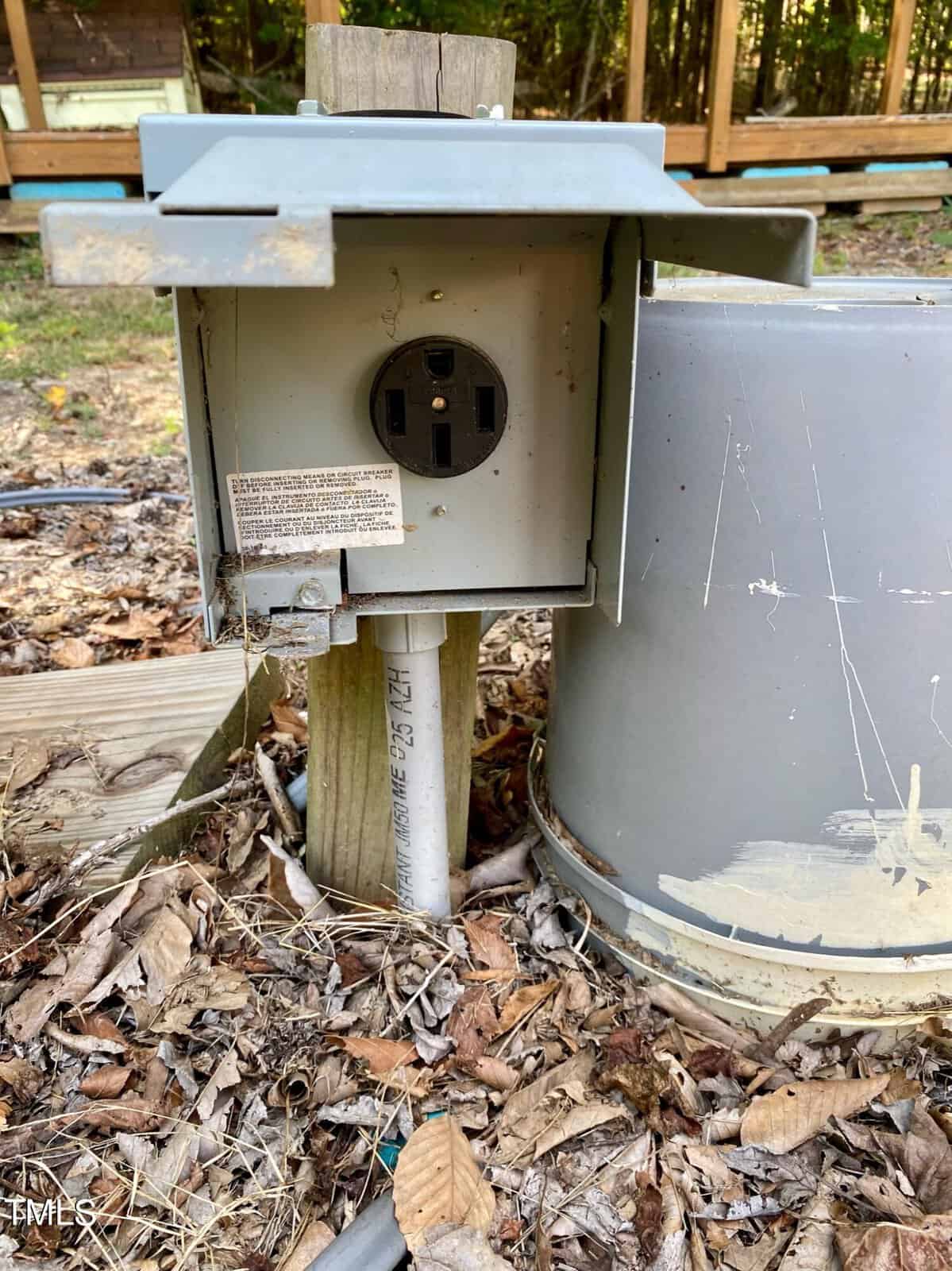 An outdoor electrical outlet with a metallic cover, mounted on a post near a large metal cylinder, surrounded by woodland debris.