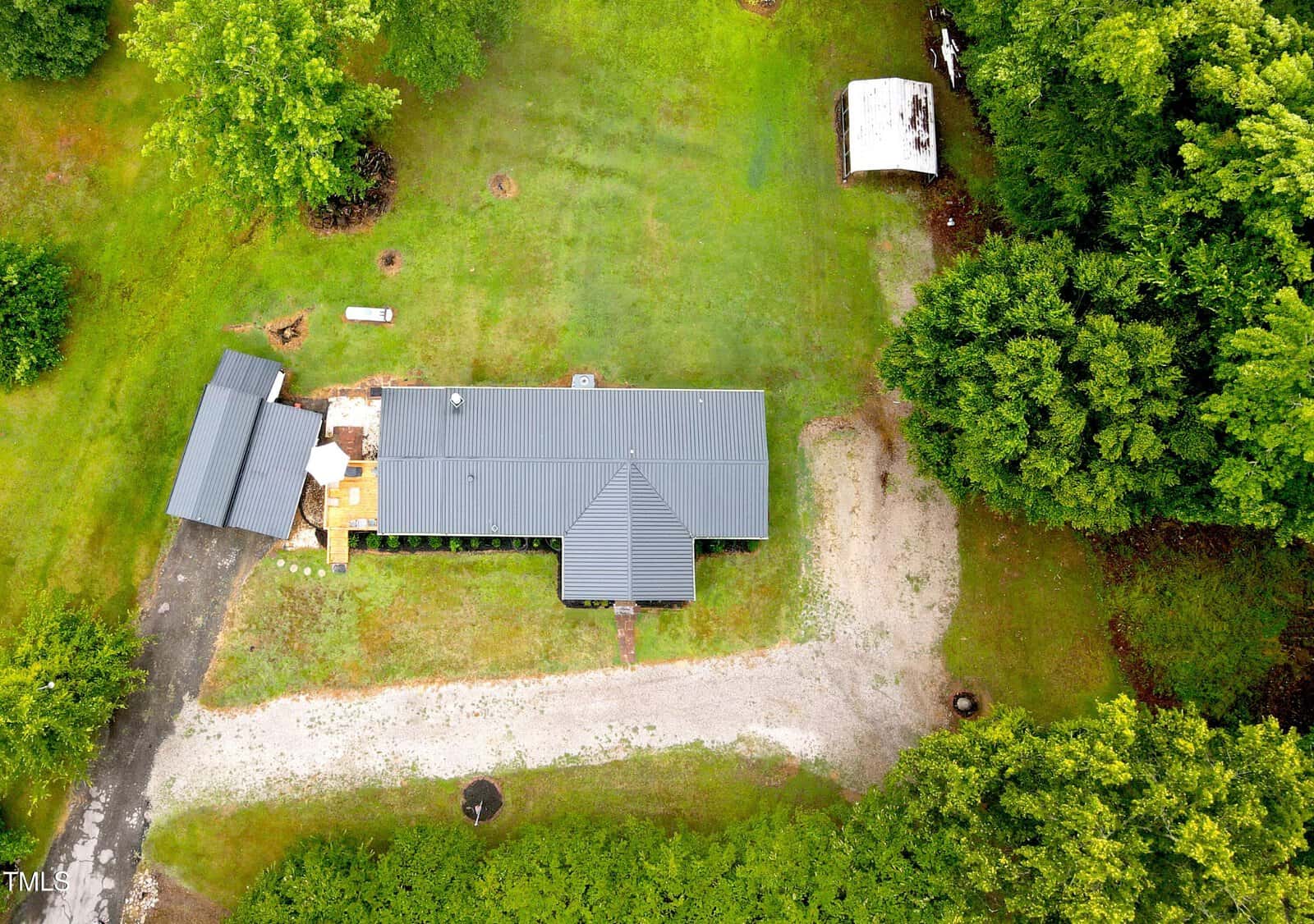 Aerial view of a rural property with a gray metal roofed house surrounded by lush green trees and a small cleared yard. there's a dirt path and a few scattered outbuildings.