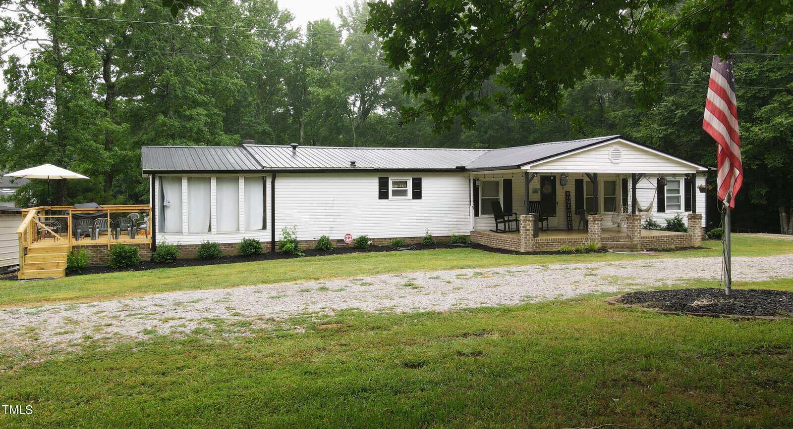 A long, white single-story home with a metal roof and a covered porch at 194 Waters Edge Road, Henderson NC. There’s an American flag on the left and a wooden deck with a sun