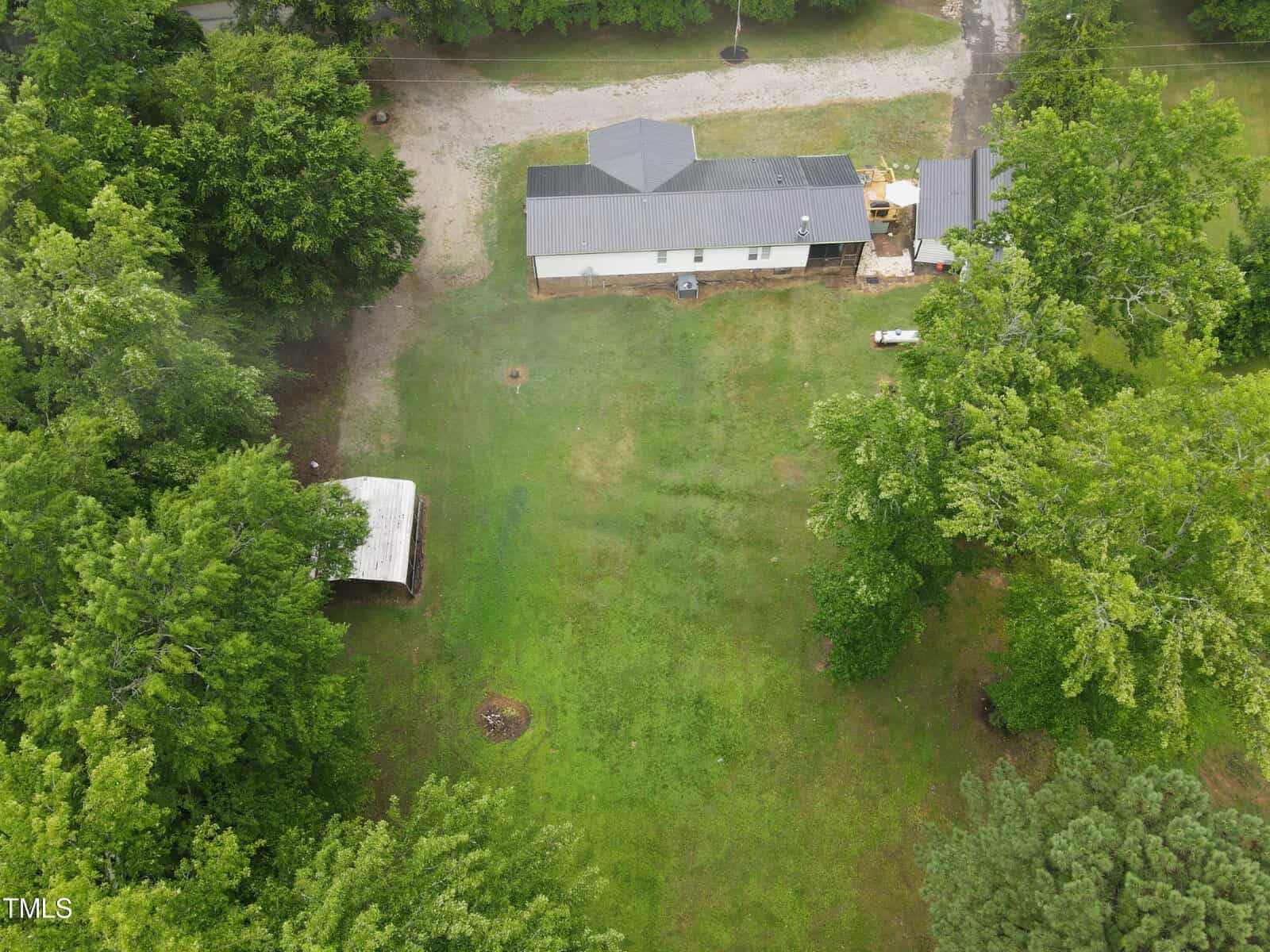 Aerial view of a residential property with a white one-story house surrounded by green trees and a large grassy yard. there is an adjacent small building or shed.