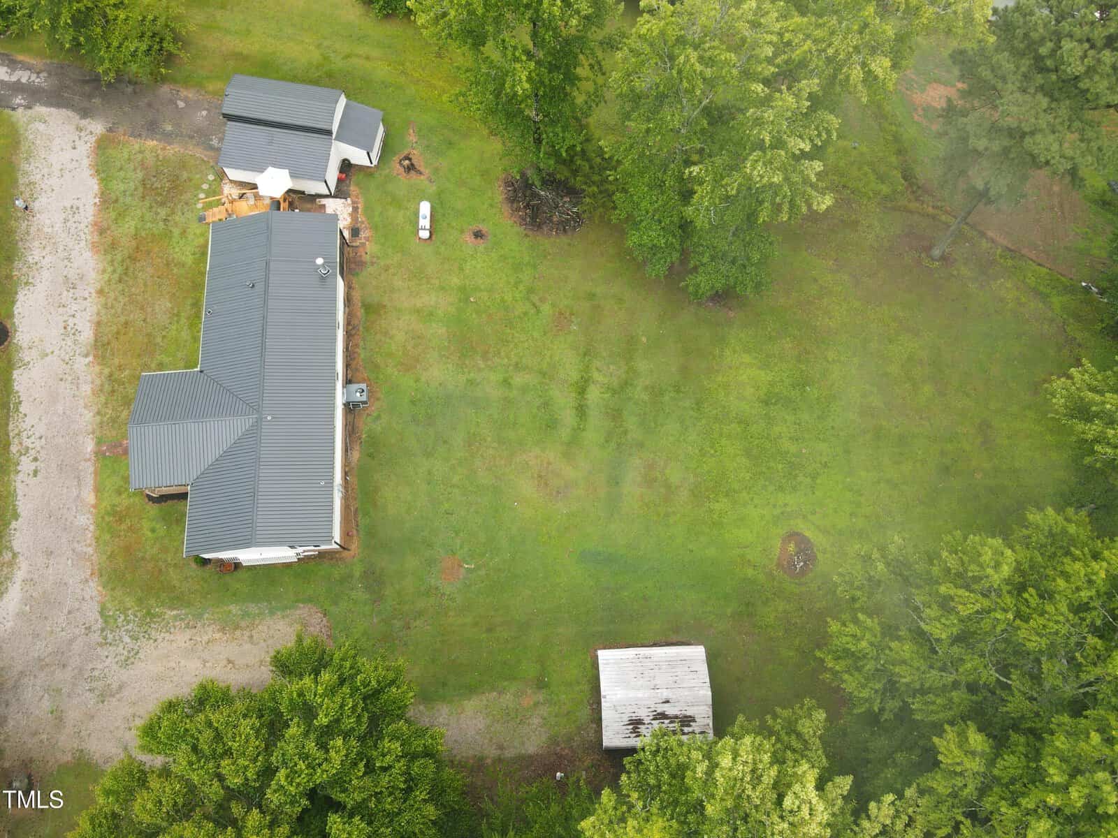 Aerial view of a rural property with two buildings featuring gray metal roofs, surrounded by green trees. a white car is parked between the buildings, near a dirt road.