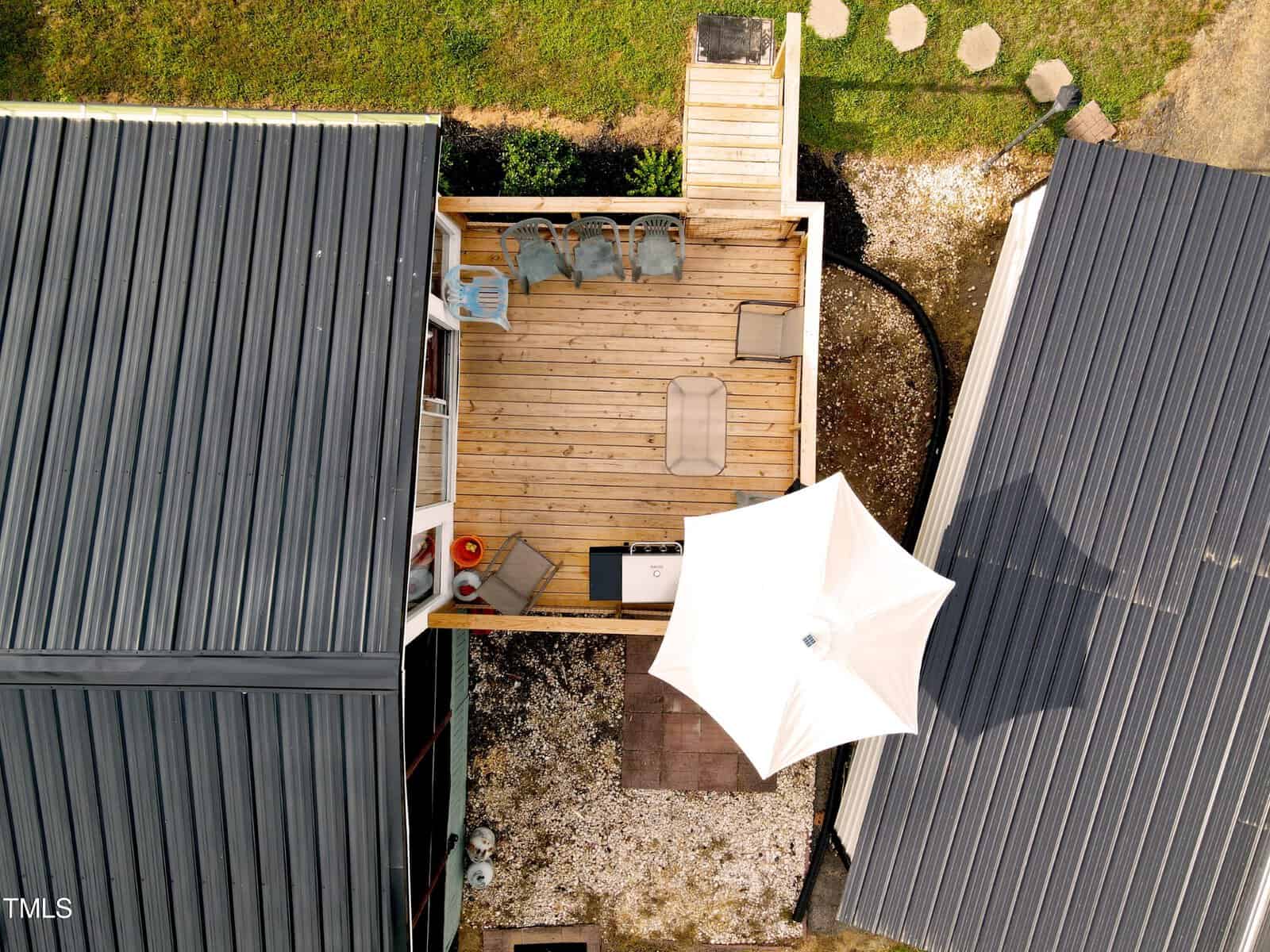 Aerial view of a small rooftop terrace with wooden decking, surrounded by two gray buildings. the terrace features a white umbrella, seating area, and a few potted plants.