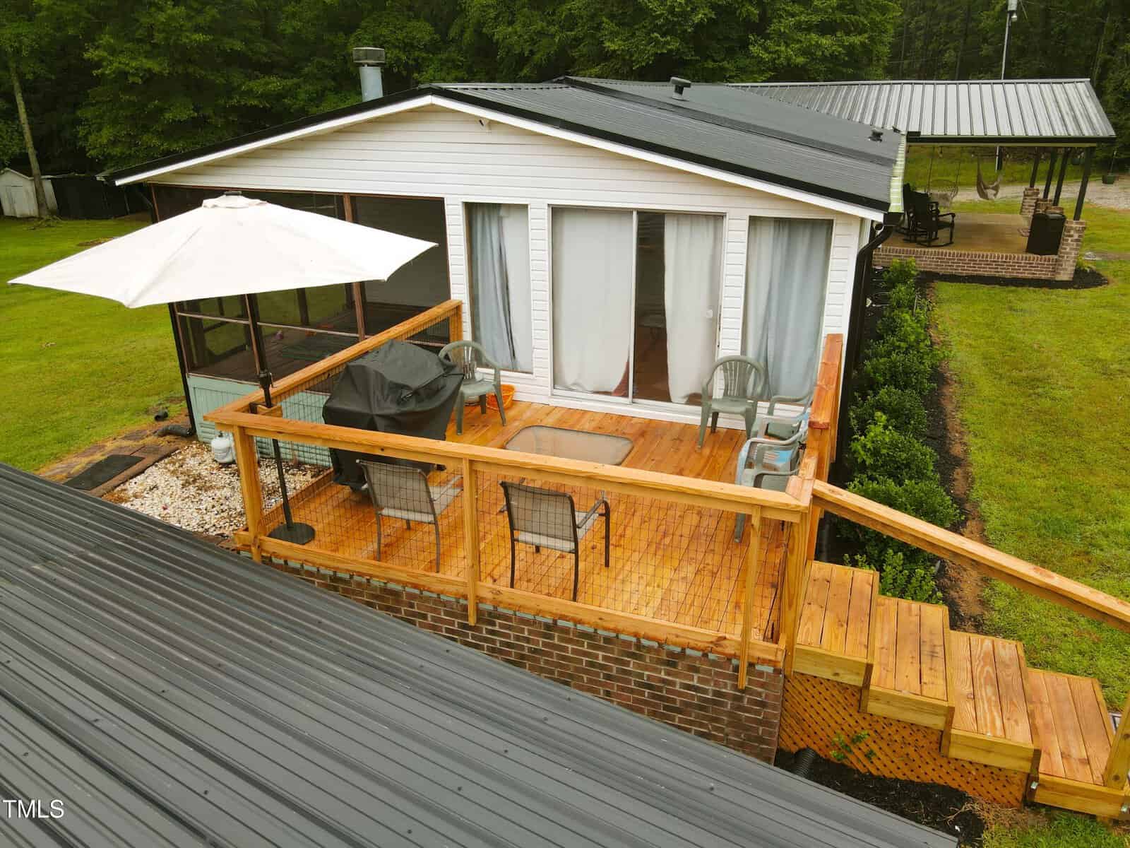 Aerial view of a backyard with a wooden deck featuring patio furniture under an open white umbrella, adjacent to a metal-roofed house. the deck includes a grill and is surrounded by lush greenery.