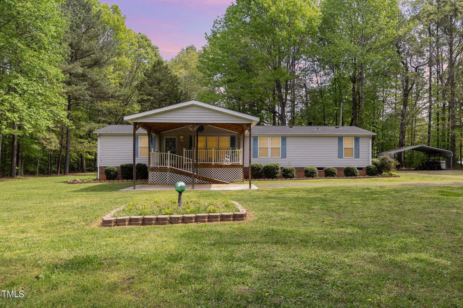 The single-story gray house at 2205 Lauren Mill Drive in Oxford features a charming covered porch, surrounded by a well-maintained lawn and trees. A gravel driveway leads to the carport on the right, all beneath a sky tinged with pink, signaling sunset or sunrise.