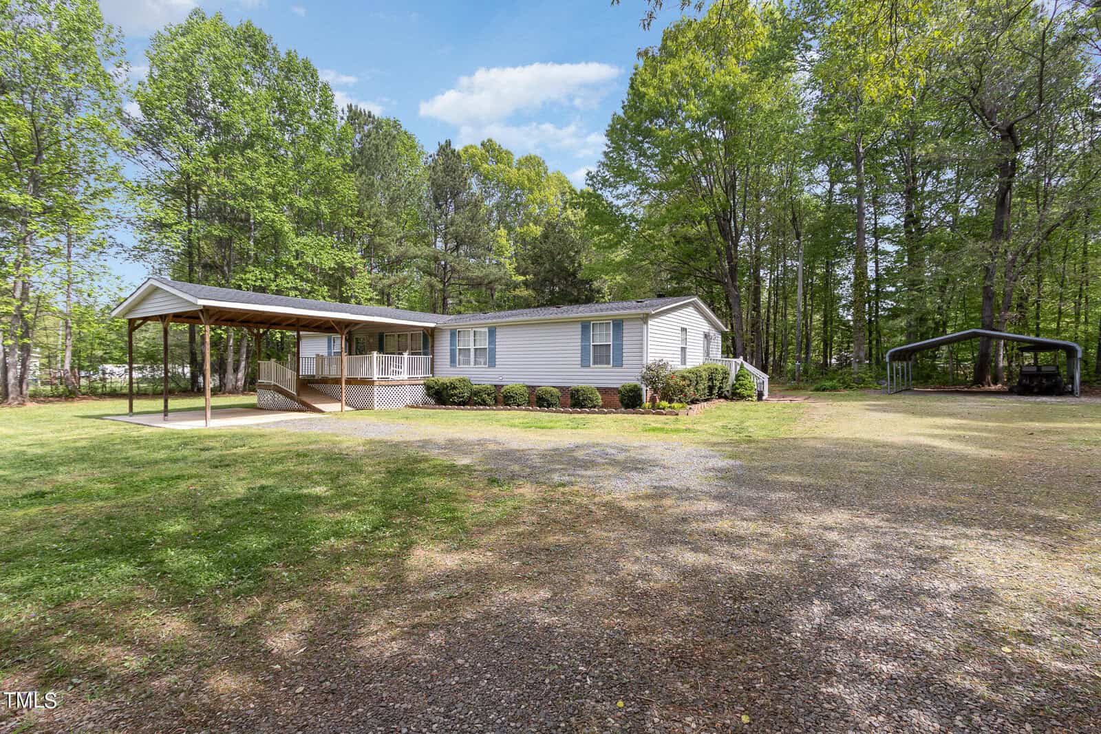 A white manufactured home with a covered porch sits amid greenery, surrounded by tall trees. A gravel driveway leads to the house, and a carport is visible to the right. The sky is clear and blue, casting natural light on the scene.
