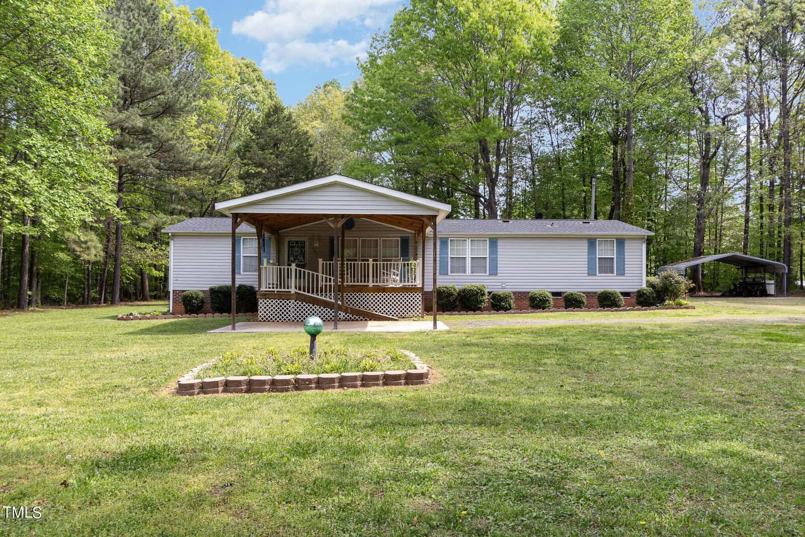 A white manufactured home with a covered porch sits amid lush greenery and trees. A small garden bed is in the front yard, and a driveway leads to a carport on the right. The sky is clear with a few clouds.