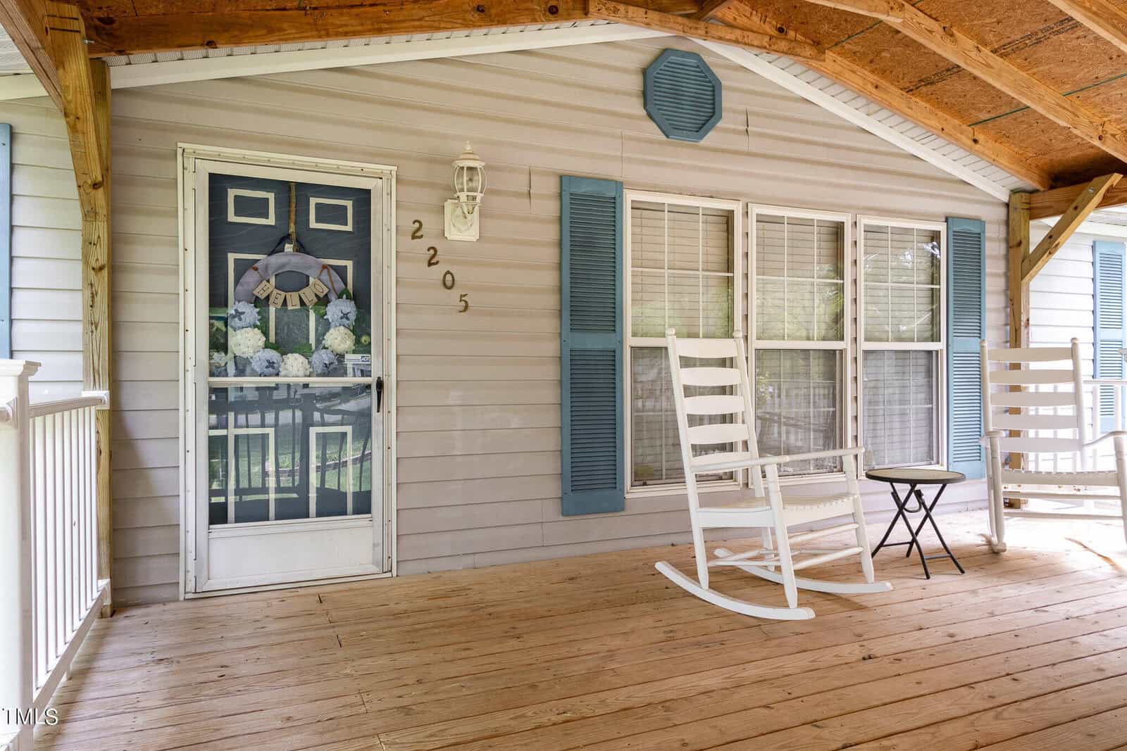 A cozy porch with two white rocking chairs and a small table on a wooden deck. The door is decorated with a floral wreath, and large windows are on the right side. The exterior is light-colored with blue shutters.