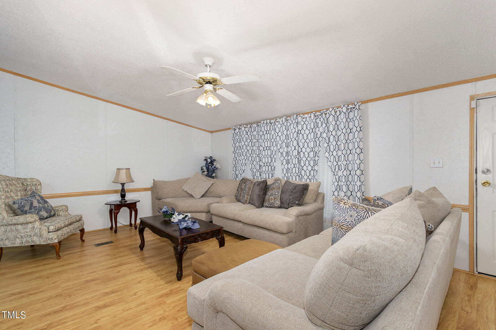 A cozy living room with beige sofas arranged around a wooden coffee table. The room features hardwood flooring, a ceiling fan with lights, patterned curtains, and a comfortable armchair. A door is visible on the right.