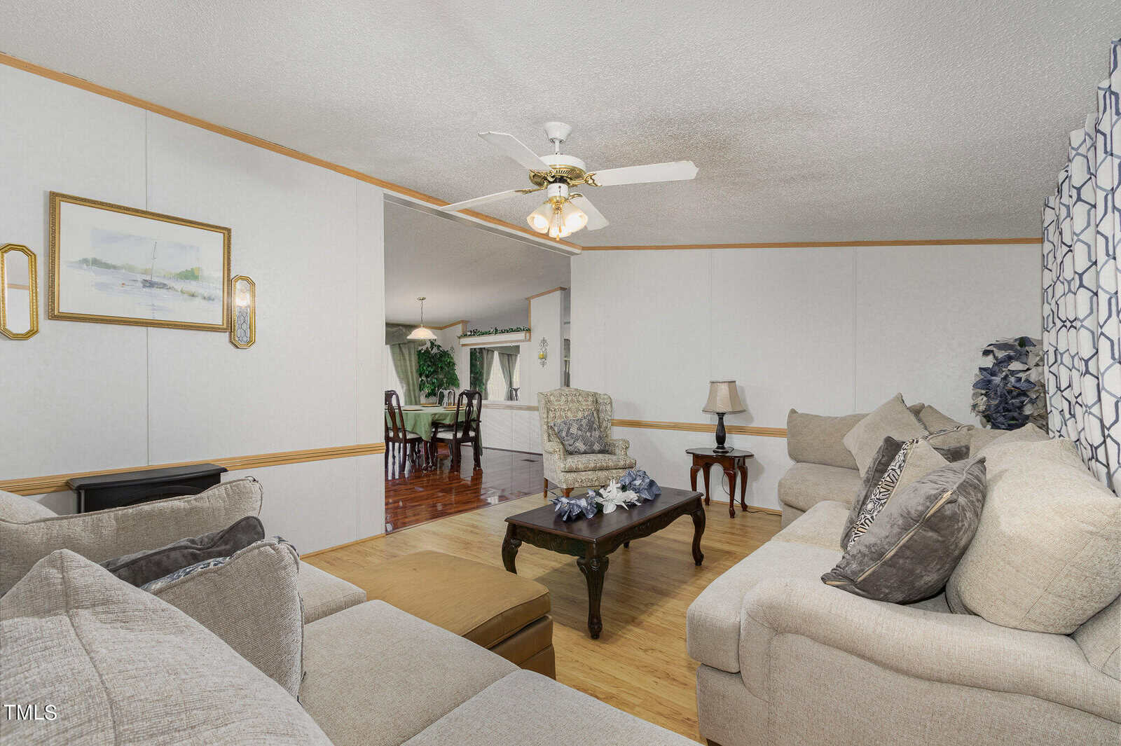 A cozy living room with beige sofas, patterned cushions, and a wooden coffee table. A ceiling fan hangs above. In the background, there's a dining area with wooden chairs and a decorative plant. The room has a light and airy ambiance.