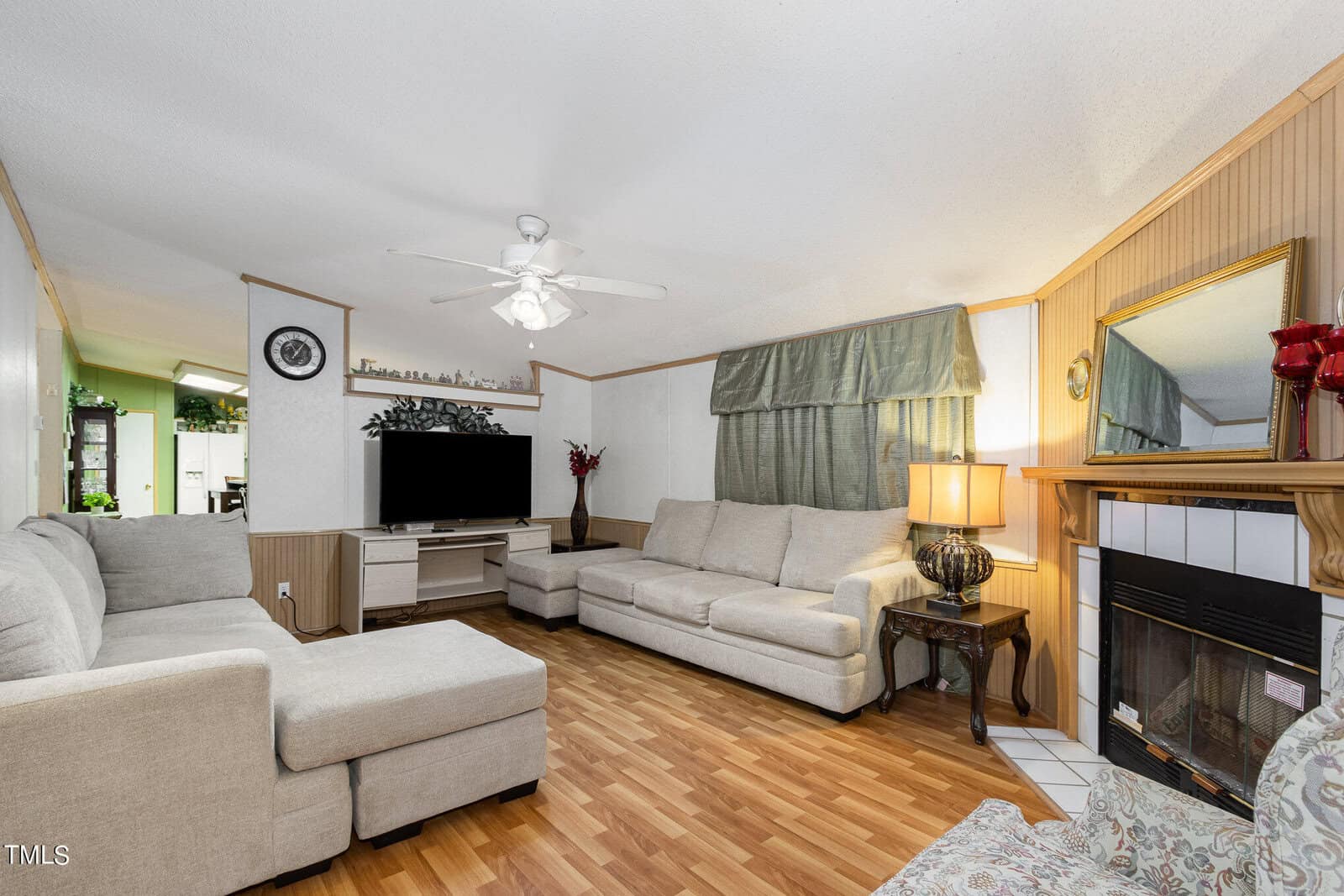 A cozy living room with light wood flooring, two beige sofas, a flat-screen TV on a stand, a ceiling fan, and a fireplace. Beside the fireplace is a table with a lamp. Green curtains adorn the window, adding a touch of color to the decor.