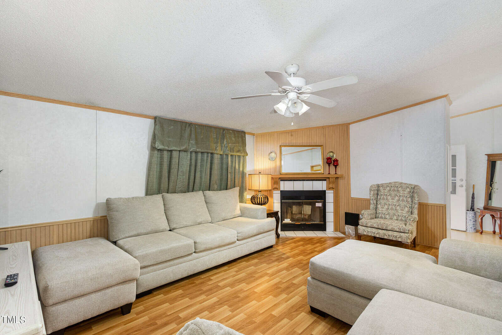 A cozy living room with beige sectional sofas and a matching ottoman. A fireplace with a mirror above is centered on a wooden accent wall. A patterned armchair and side table with a lamp sit nearby. Hardwood floors and a ceiling fan complete the room.