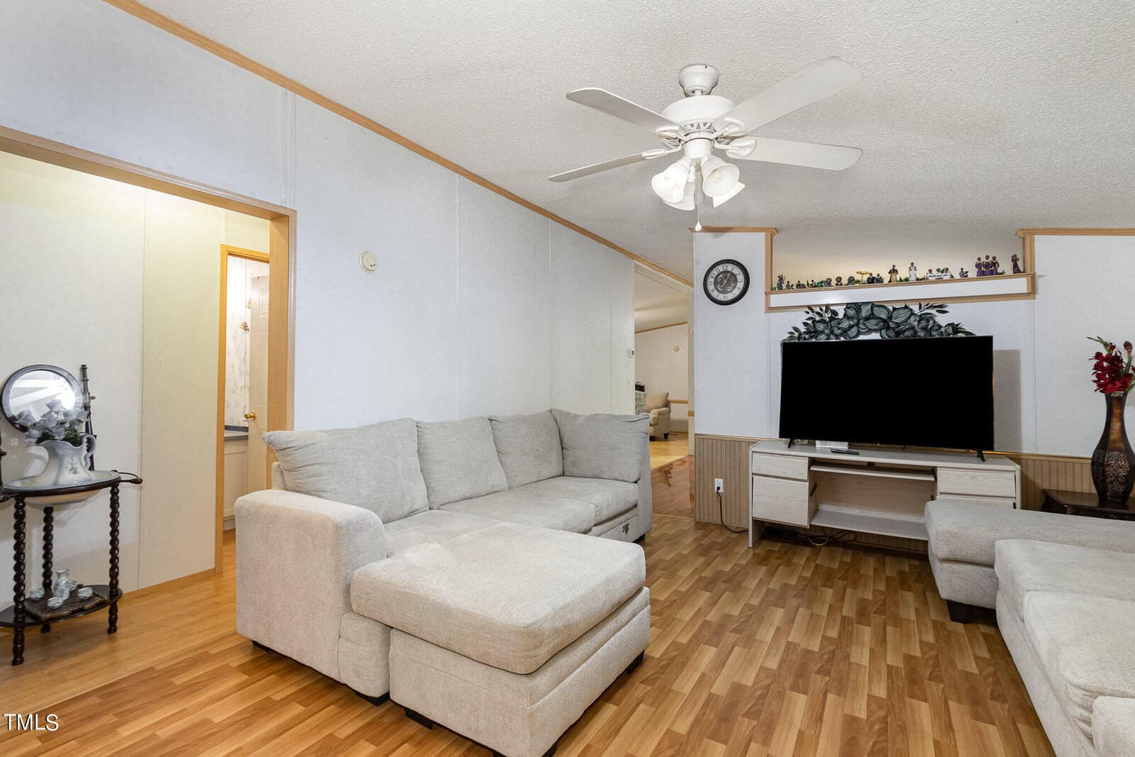 A cozy living room with light wood flooring, featuring a beige sectional sofa and a ceiling fan. A large flat-screen TV sits on a white media console, with decorative items on a shelf above. A small, round table with a decorative piece is near an open door.