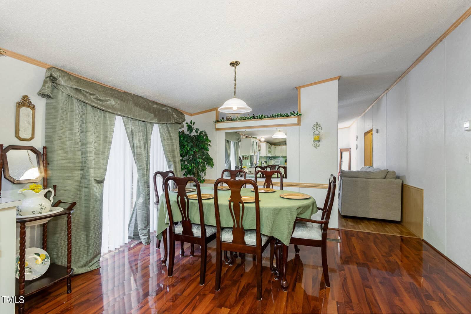 Dining room with a wooden table and six chairs, green tablecloth, pendant light, and decorative plants. A cabinet with a mirror and lamp is on the left. Open view to a kitchen and a living area with a sofa in the background.