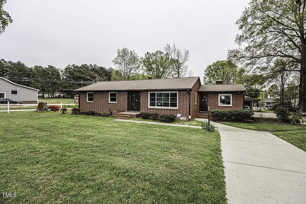 A single-story brick house with multiple windows and a white door, surrounded by a well-manicured lawn, trees, and a clear sky. a concrete pathway leads to the entrance.