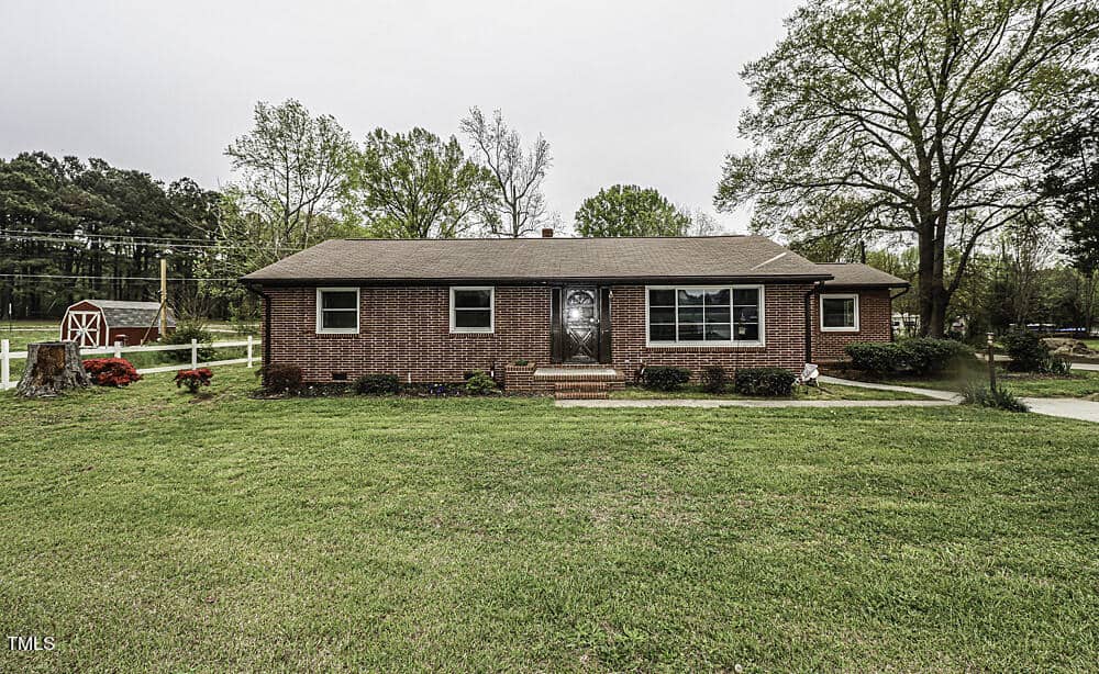 A single-level brick suburban house with a dark shingled roof, surrounded by a well-maintained green lawn with small flowering bushes near the foundation. a wooden fence and trees are visible in the background.