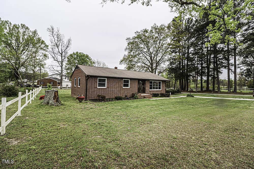 A small, single-story brick house with a white picket fence, surrounded by a large, grassy yard with several tall trees in a rural setting.