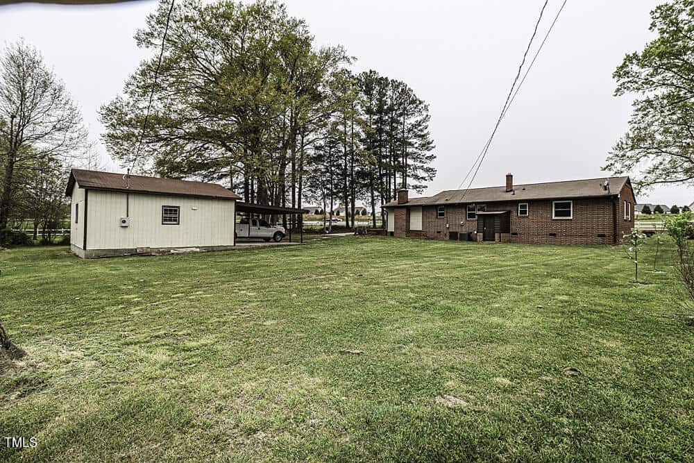 A spacious backyard featuring a brick house with an adjacent beige outbuilding and a covered carport. a large green lawn with scattered trees under a cloudy sky.