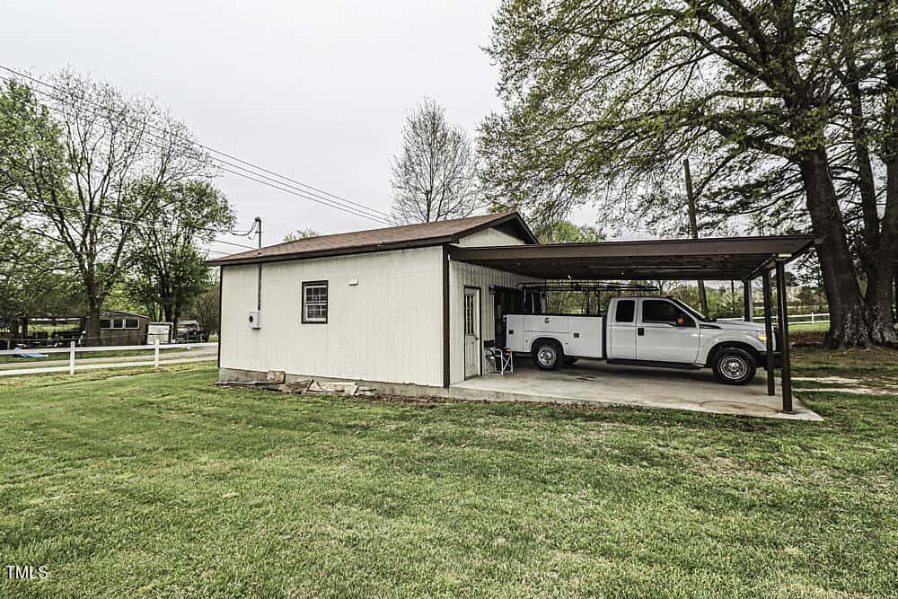 A white metal building with an attached carport sheltering a white pickup truck, set on a green lawn with trees and a white fence in the background.