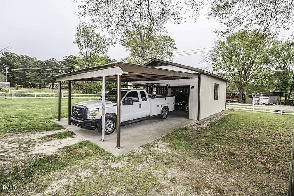A white pickup truck parked under a metal carport attached to a small workshop in a rural setting with trees and a fence visible in the background.