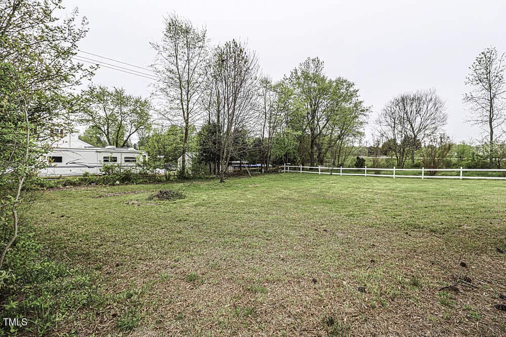 Spacious rural landscape featuring a grassy field enclosed by a white fence, with trees in the background and a camper van parked near the fence on the left.