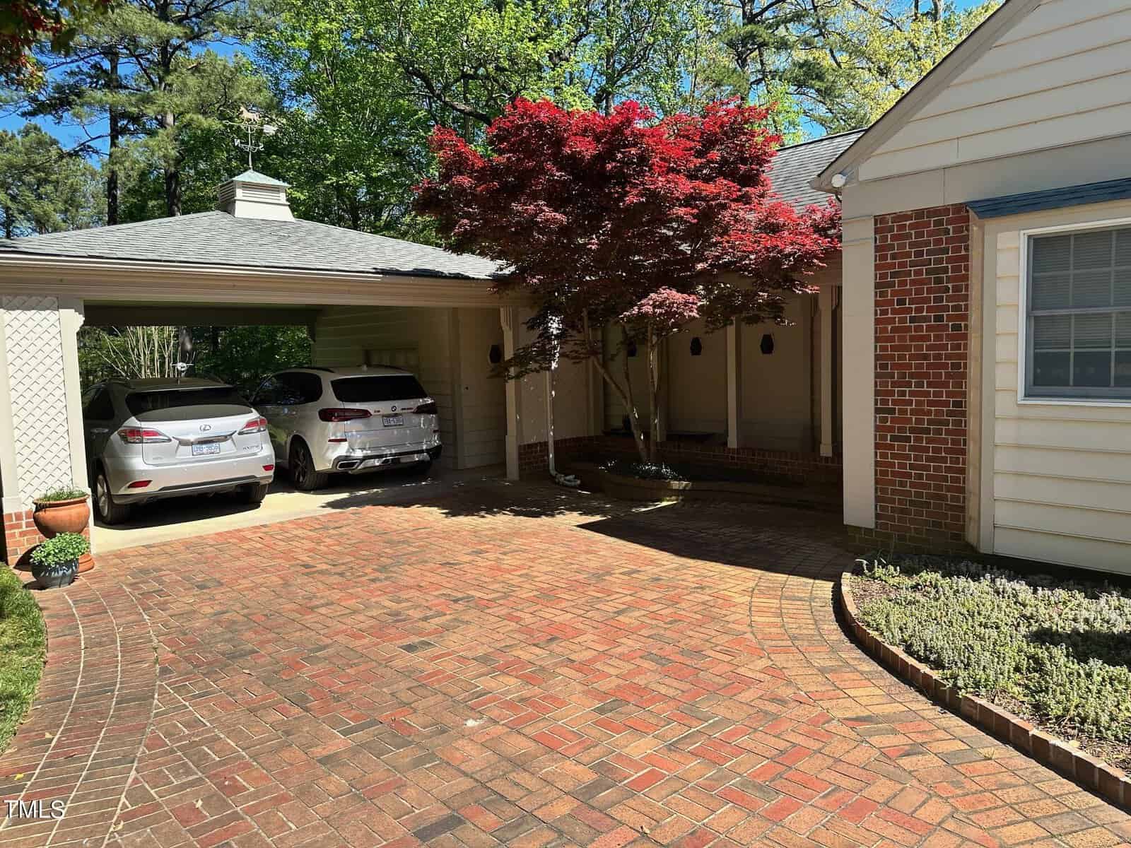 Residential driveway with two cars parked under a carport beside a brick house, featuring a vibrant red tree and well-maintained green shrubs under a sunny sky.