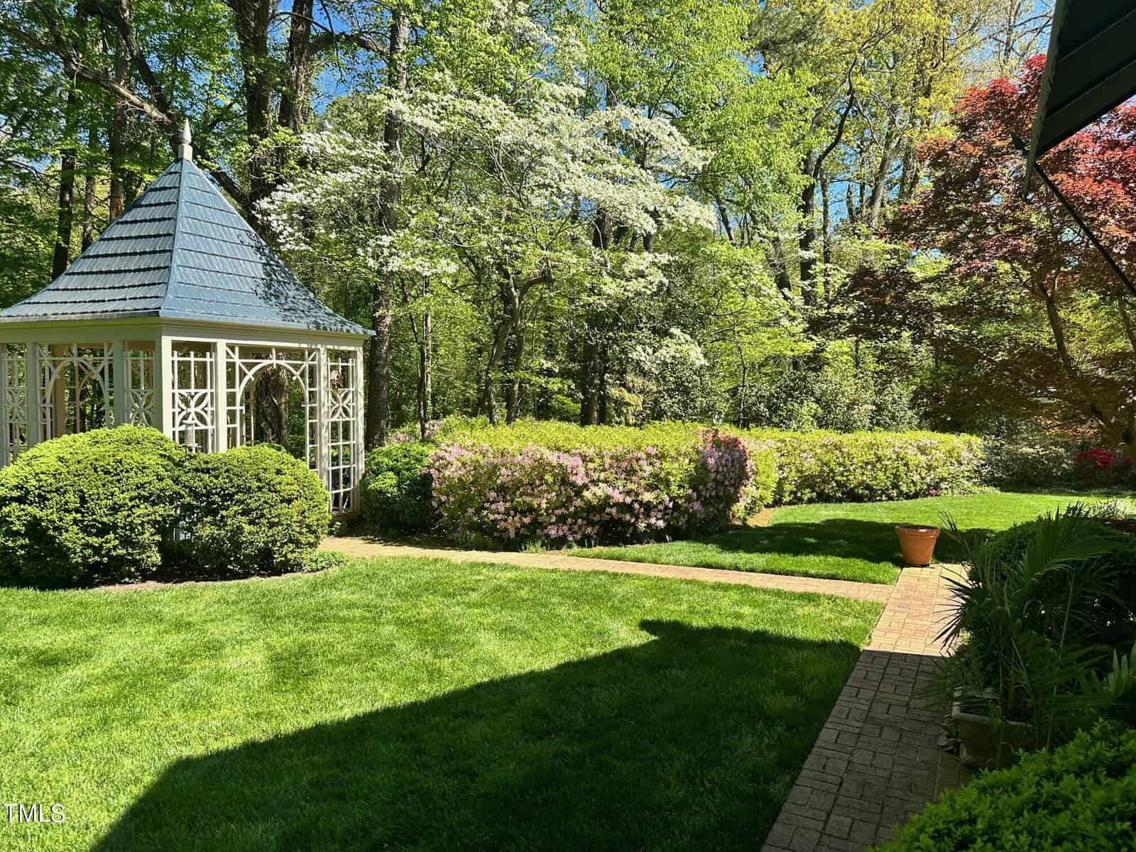A lush garden featuring a gazebo, vibrant flowering bushes, trimmed hedges, and a neatly maintained lawn, encircled by tall, leafy trees under a bright blue sky.
