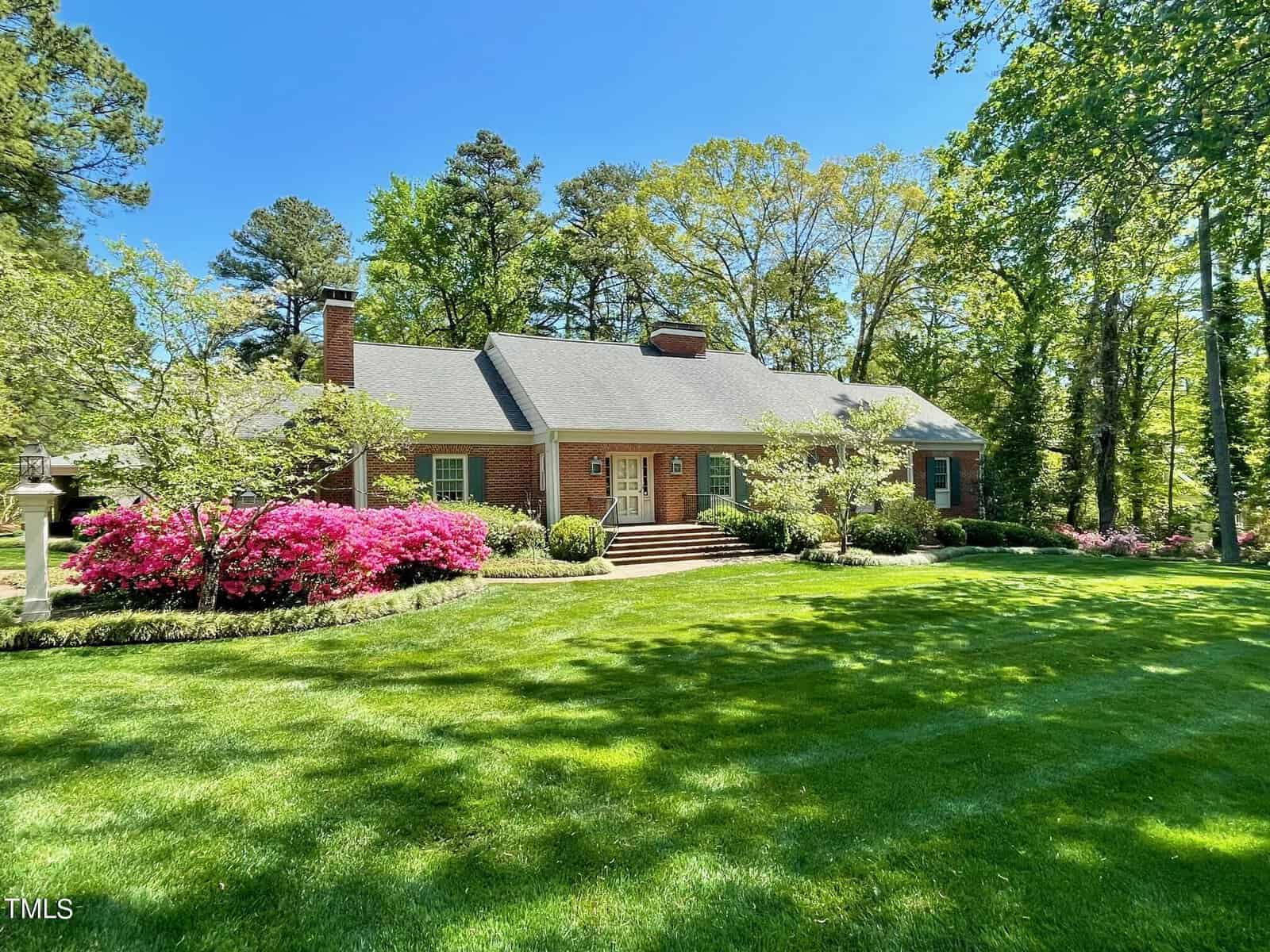 Single-story brick house at 1901 Summitt Road, Henderson NC, with a shingled roof surrounded by a lush, well-maintained lawn and blooming pink azaleas, under