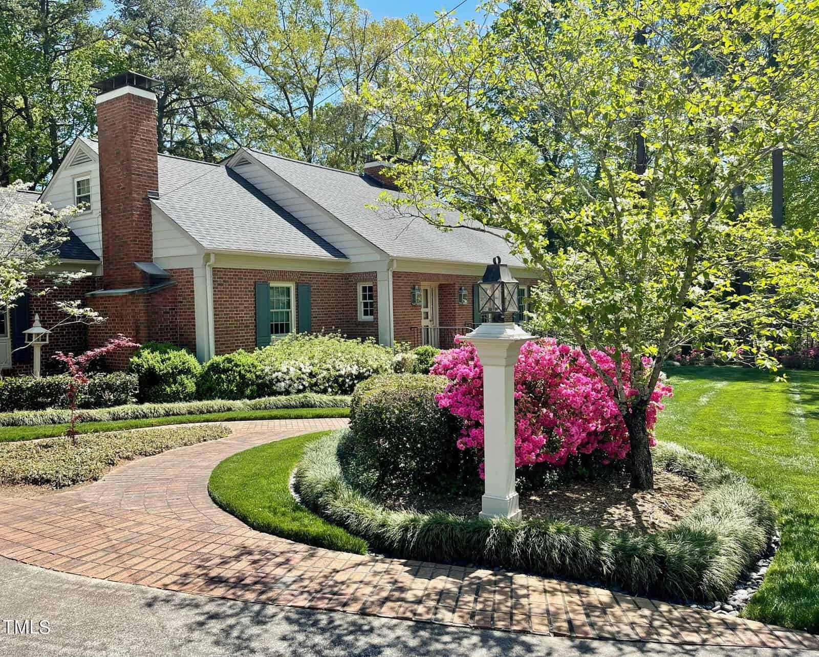 A charming brick house with a well-manicured garden featuring blooming pink azaleas, lush green bushes, and a curving brick pathway under a clear blue sky.