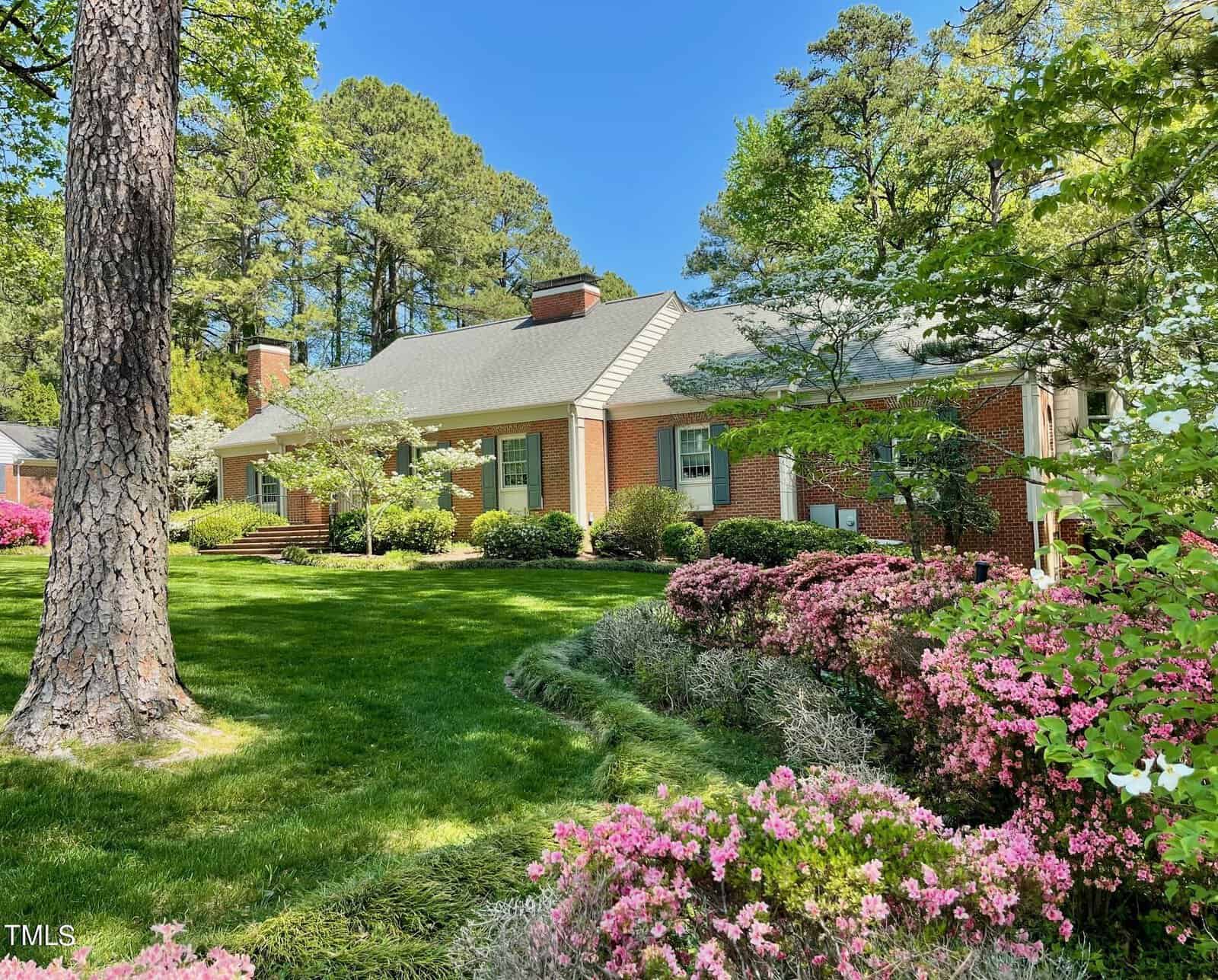 A charming brick house surrounded by lush green lawn, flowering pink shrubs, and tall pine trees under a clear blue sky.