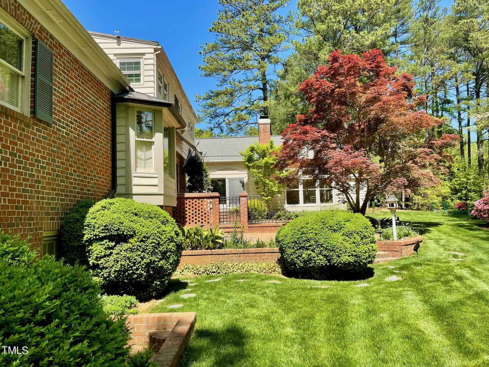 A sunny suburban landscape showcasing a well-manicured lawn and a variety of trees, including a prominent red maple beside a brick house with a partial view of a wooden arbor.