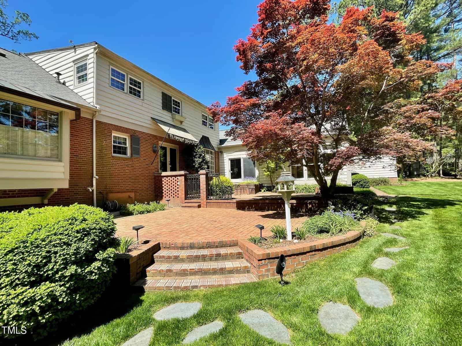 A well-manicured suburban yard featuring a brick pathway leading to a two-story house, bordered by a lush lawn, various bushes, and a striking red-leafed tree under a clear blue sky.