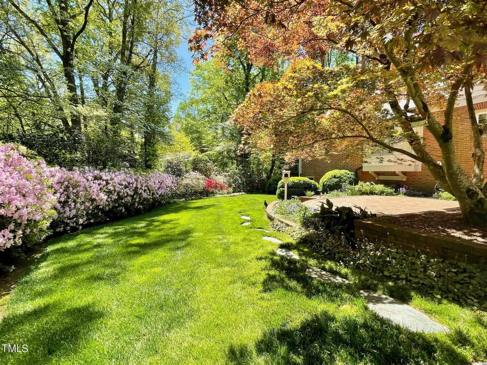 A lush garden with vibrant green grass, bordered by colorful azalea bushes and mature trees, with a brick house partially visible under a canopy of leaves.