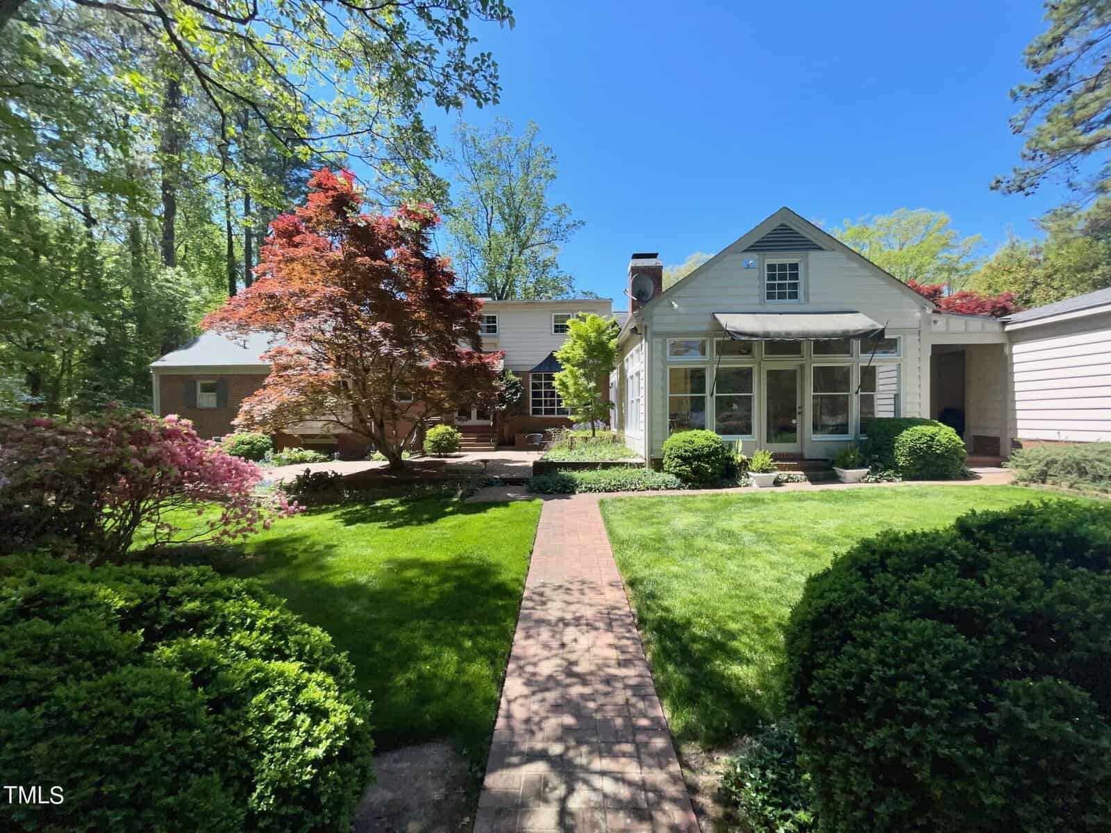 A quaint house with a beige exterior surrounded by lush greenery and colorful trees under a clear blue sky. a neat garden path leads to the front door.