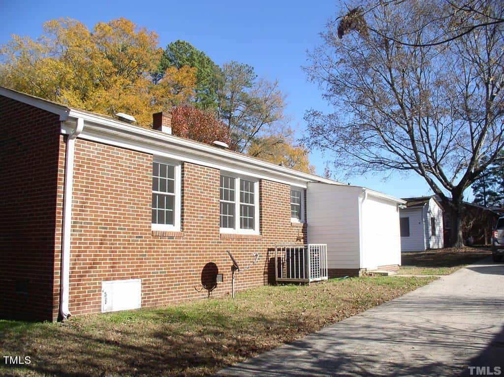 A one-story brick house with white trim, featuring a small porch and several windows. trees with autumn foliage are visible behind, under a clear blue sky.