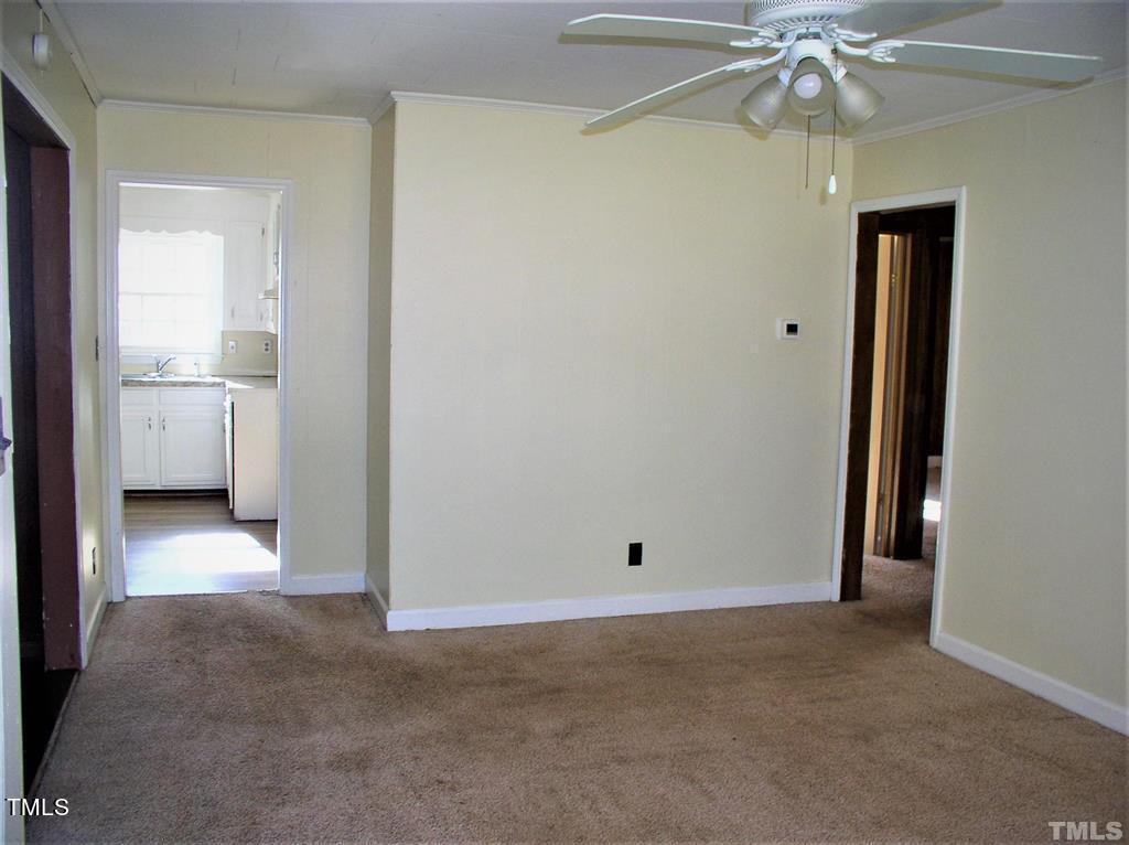 An empty room featuring beige walls and brown carpet, with an open doorway leading to a kitchen and another to a hallway, equipped with a ceiling fan.