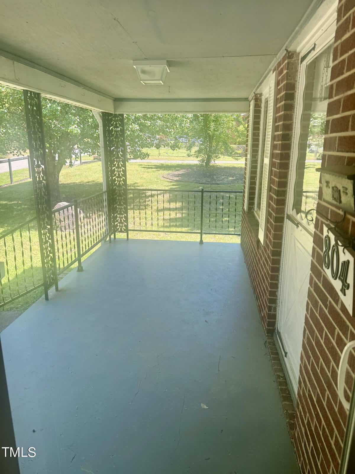 Sunlit covered porch with metal railings and brick walls, overlooking a green yard. the floor is painted blue, and shadows from surrounding trees create patterns on the surface.