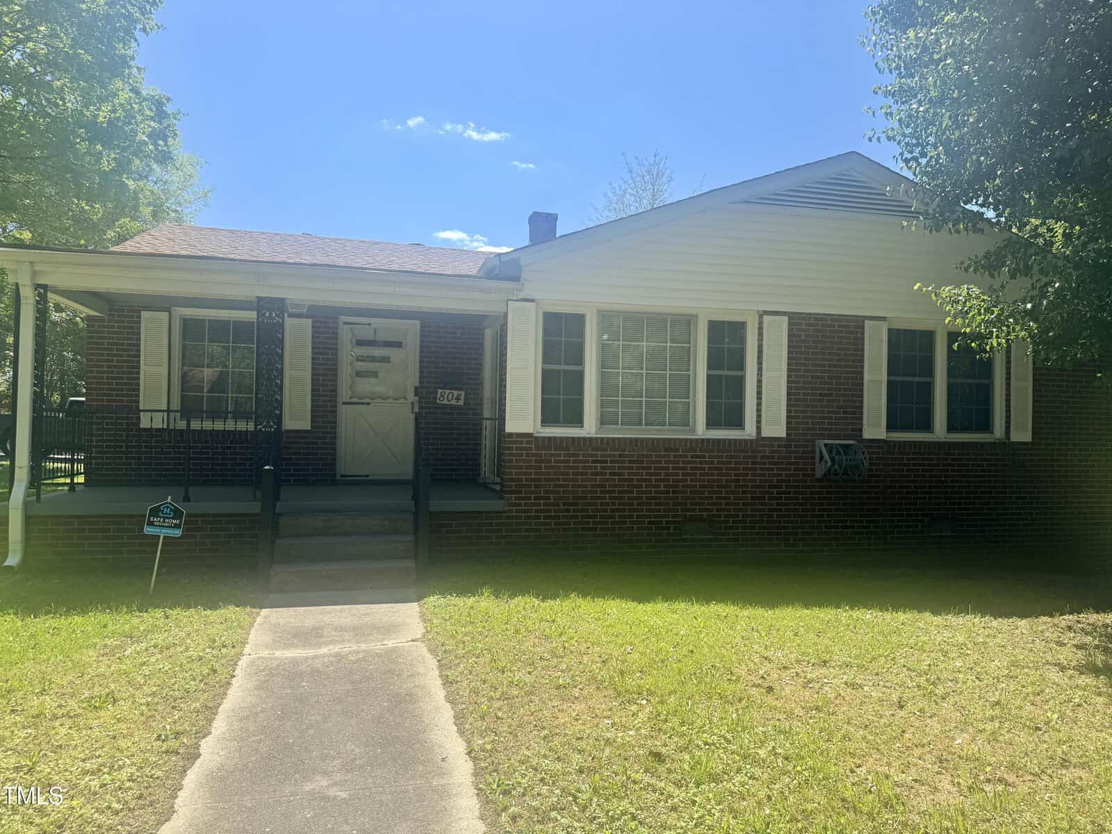 A one-story brick house with white siding and a covered entrance, located at 804 Eastside Drive, Henderson NC, featuring a small lawn and trees in the background on a sunny day. There is