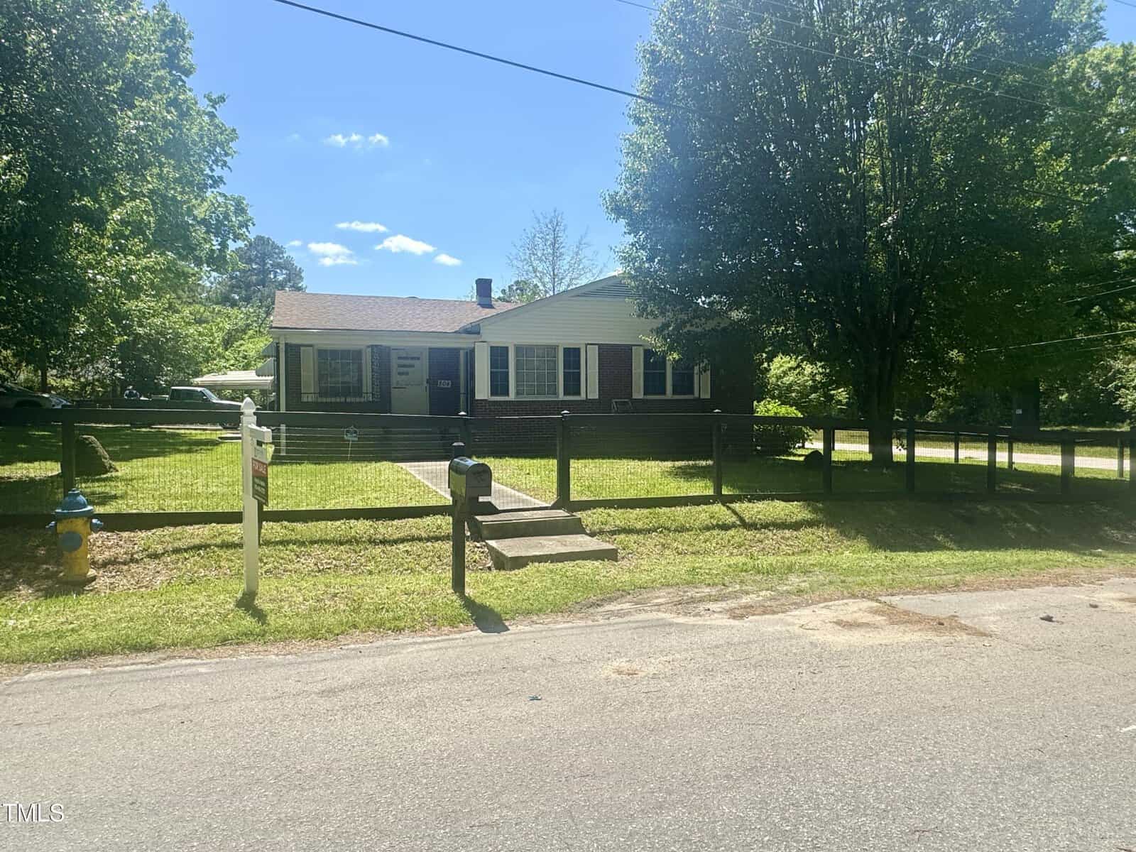 A single-story brick house with white shutters and a gray roof, surrounded by a chain-link fence, on a sunny day with lush green trees in the background.