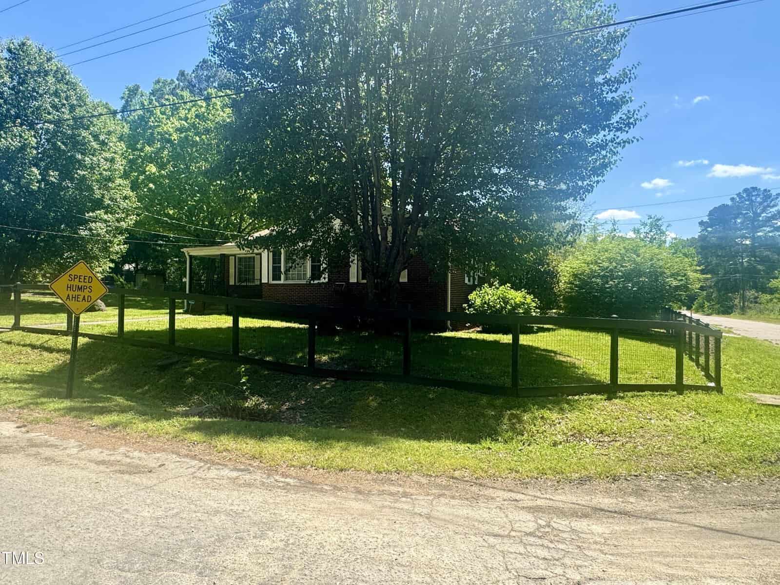 A sunny view of a suburban home with a large, leafy tree in the front yard enclosed by a low, wooden fence. a road with a "speed hump ahead" sign is visible on the left.