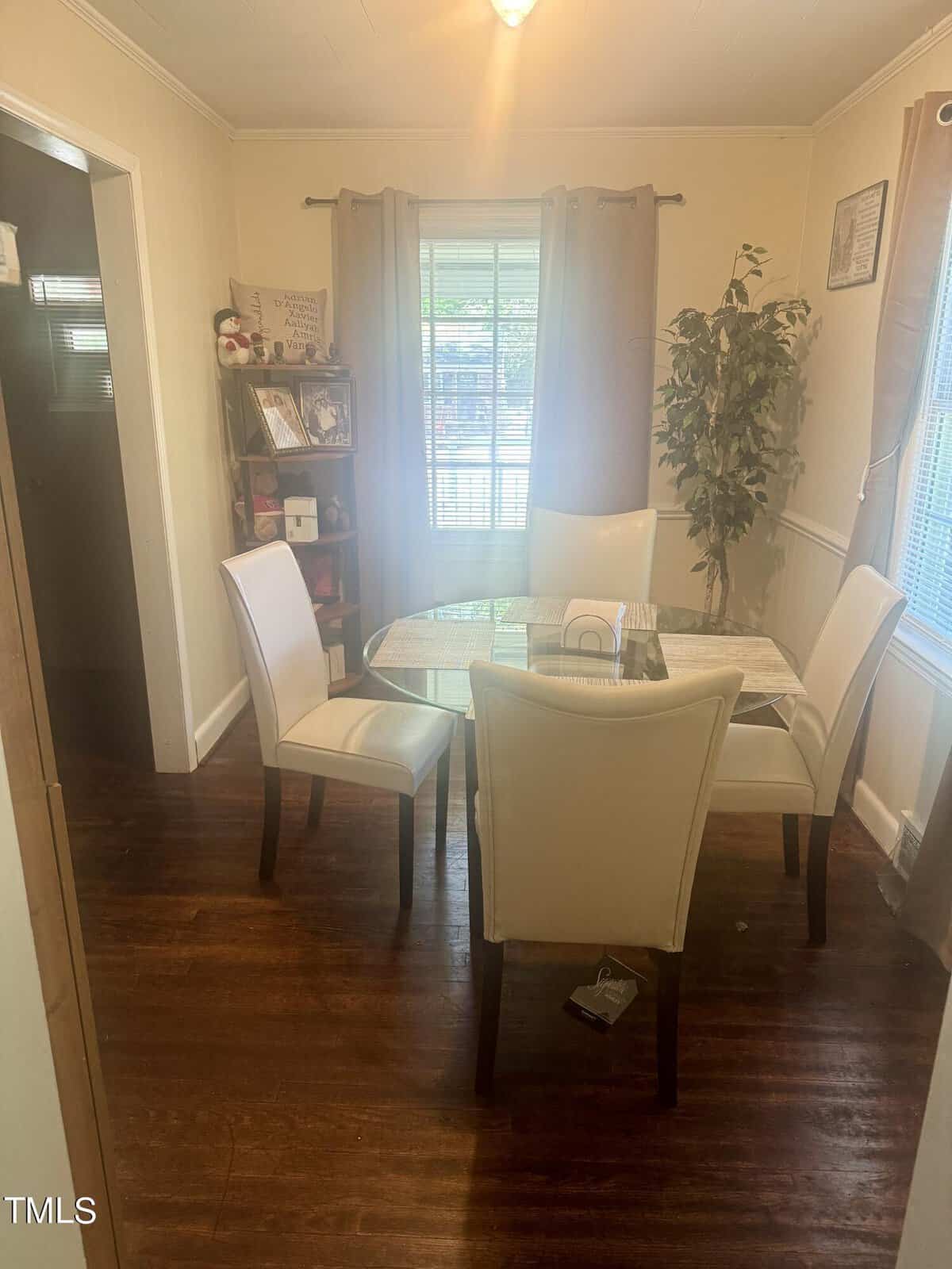A cozy dining room with a round white table and four white chairs on a wooden floor, a plant in the corner, and a bookshelf by the window with curtains.