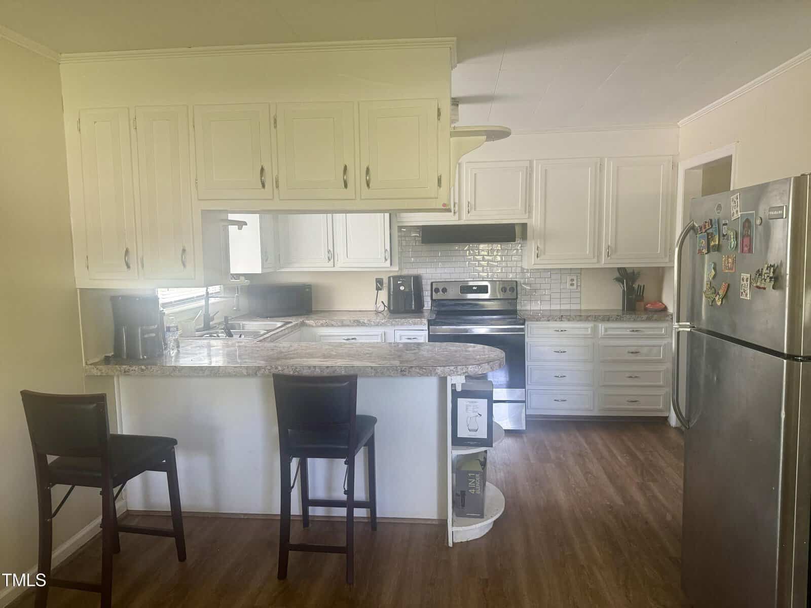 A modern kitchen with white cabinetry and stainless steel appliances. there's a granite countertop with bar stools, a fridge covered in magnets, and a sunlight-filled interior.