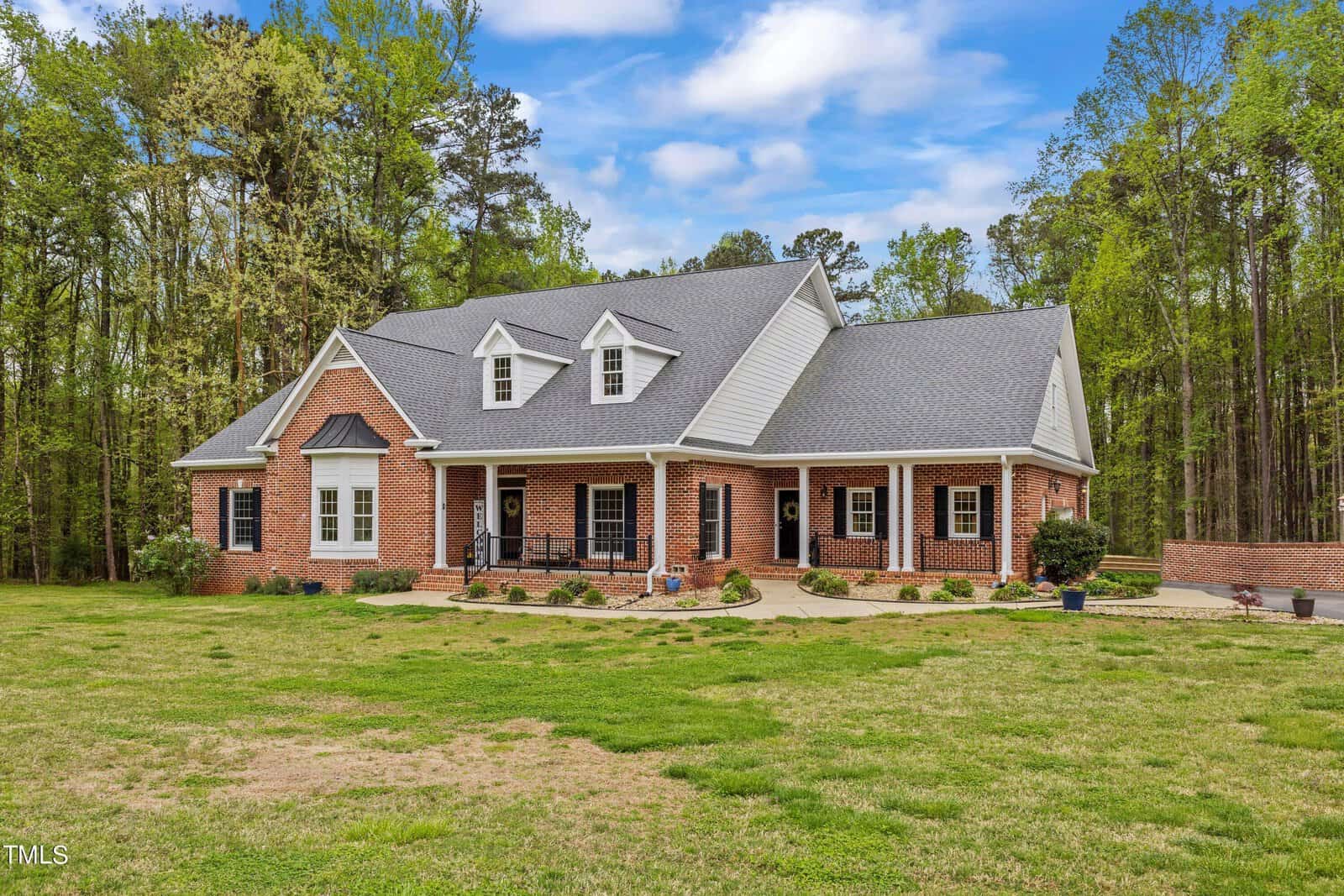 Nestled at 4050 Hugh Davis Road, Youngsville, this large brick house boasts a gray roof and is surrounded by lush green trees and a grassy lawn. The home features a charming front porch with columns and several dormer windows on the roof, all under a partly cloudy sky.