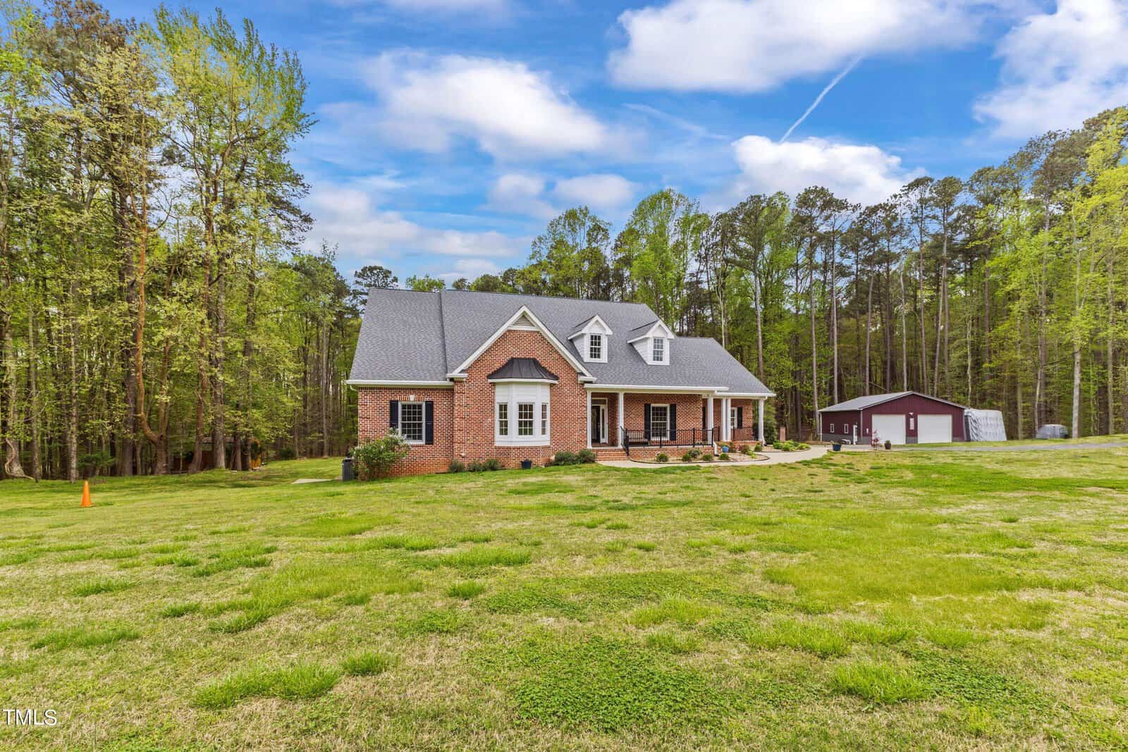 A brick house with a gray roof is situated on a grassy lawn surrounded by trees. A small building is visible in the background under a partly cloudy sky.